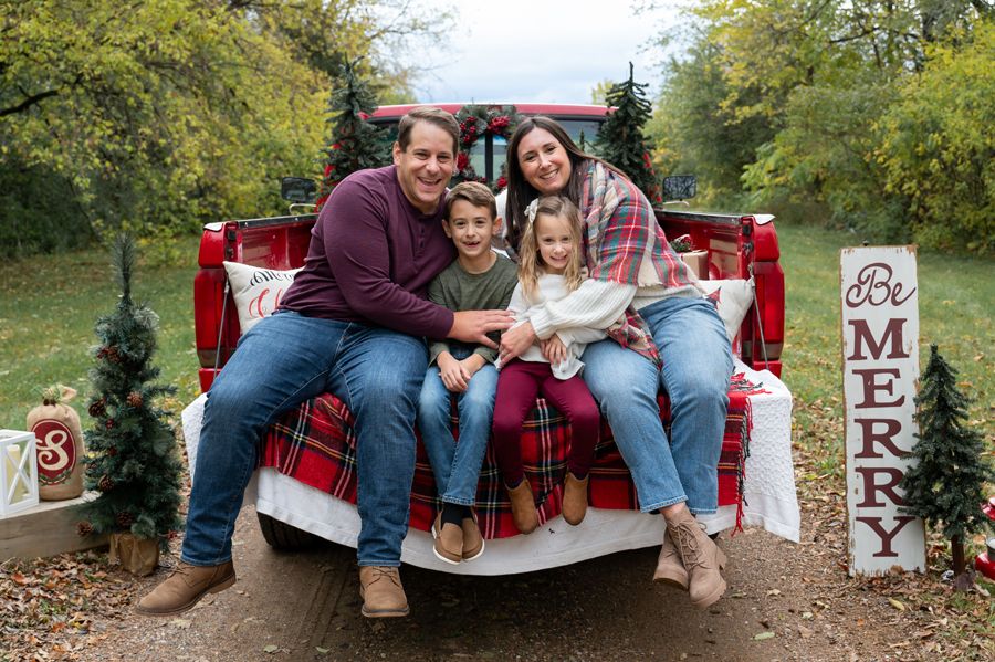A family sits together in the bed of a red pickup truck decorated for Christmas photos.