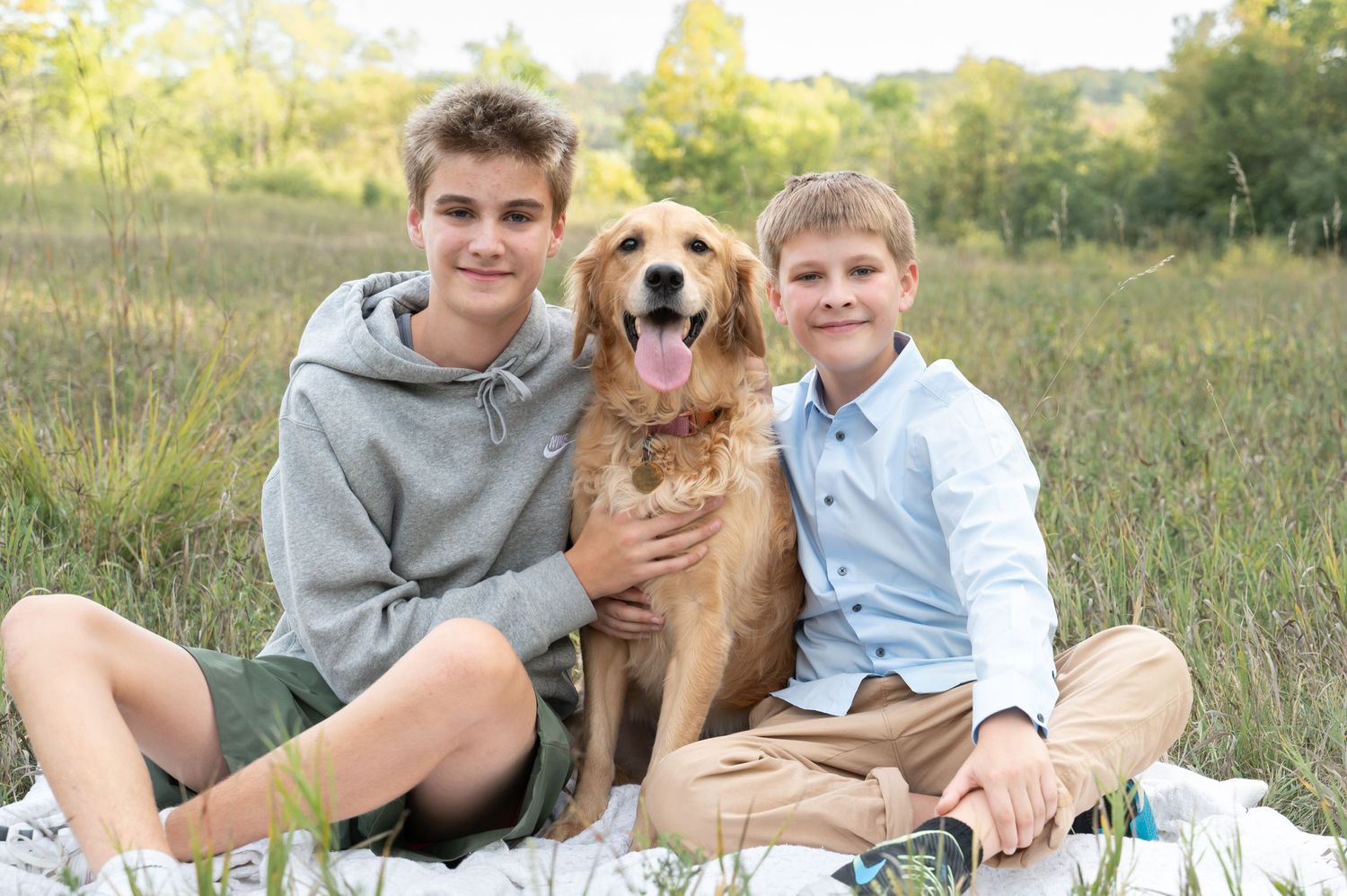 Two boys sitting outdoors with golden retriever dog on blanket in field.