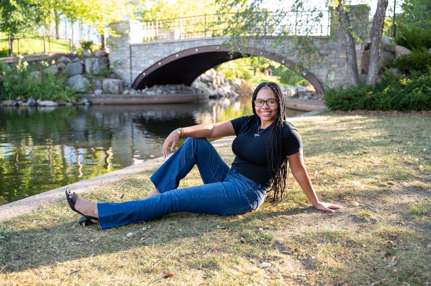 Young woman in black top and jeans sitting on grass by a scenic stone bridge and pond in a park.