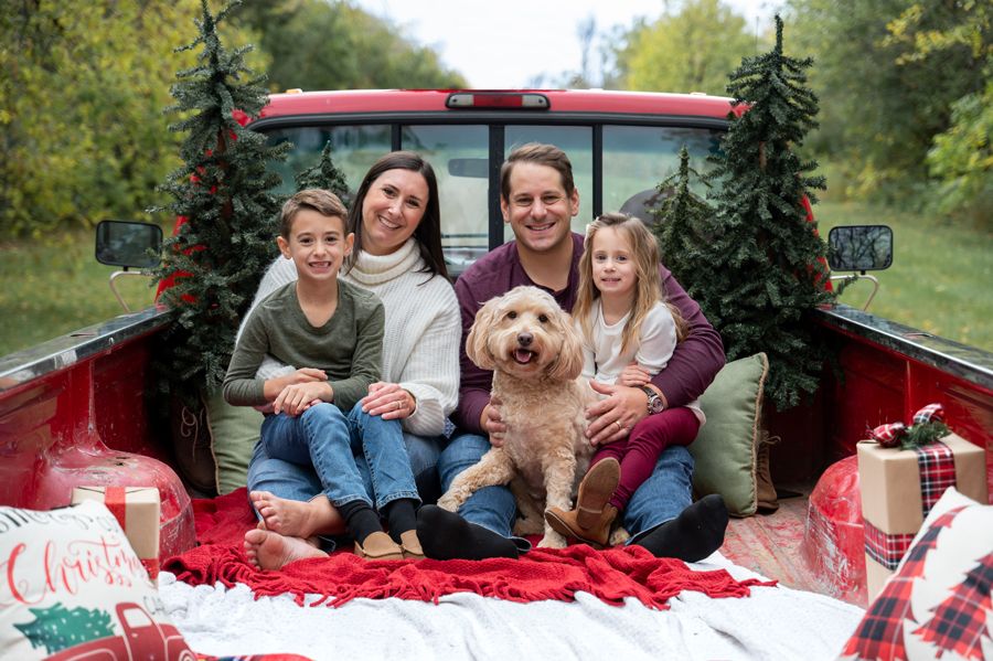 A family and their golden retriever sit together on a red blanket in a festive truck bed setting.