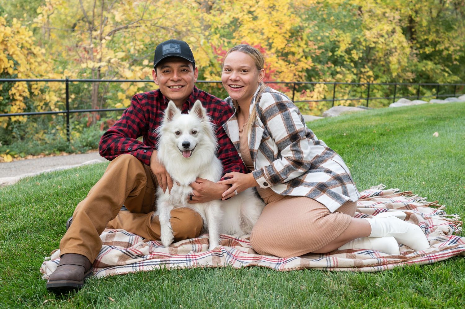 Couple in plaid shirts sitting on blanket with white fluffy dog during autumn photo session.