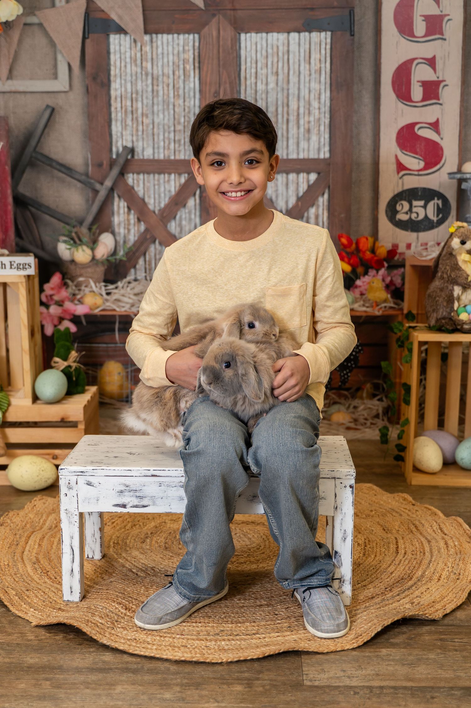 A young person sits on a white bench holding two gray rabbits in a rustic Easter-themed setting with decorative eggs and flowers.