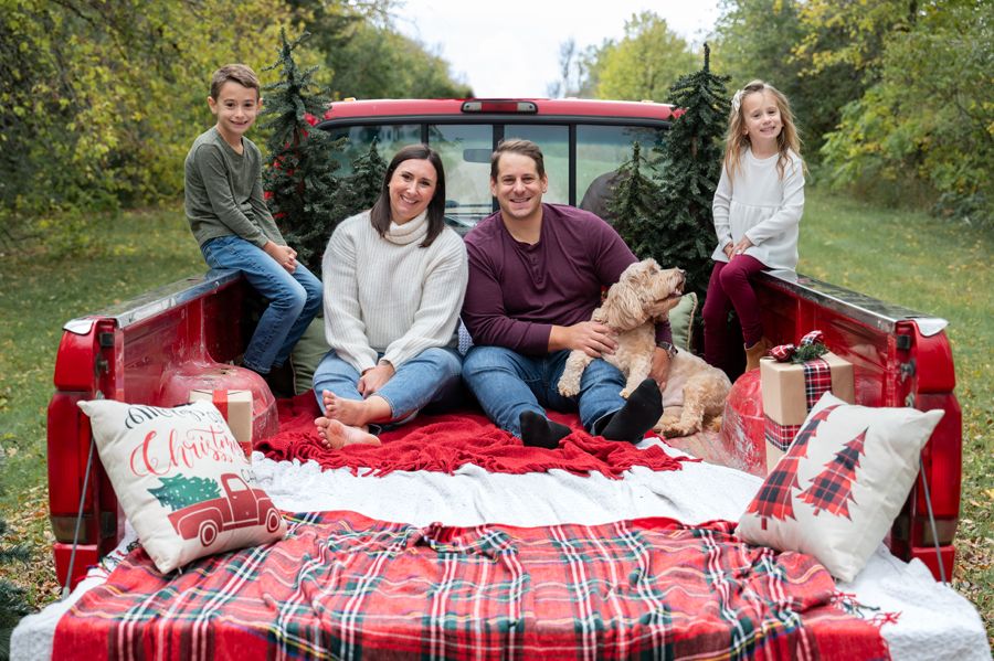 A family with children and a dog pose on a red plaid blanket in a pickup truck bed.