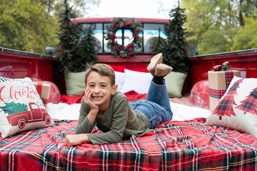 A child poses lying down on a plaid blanket in a truck bed with Christmas decorations.