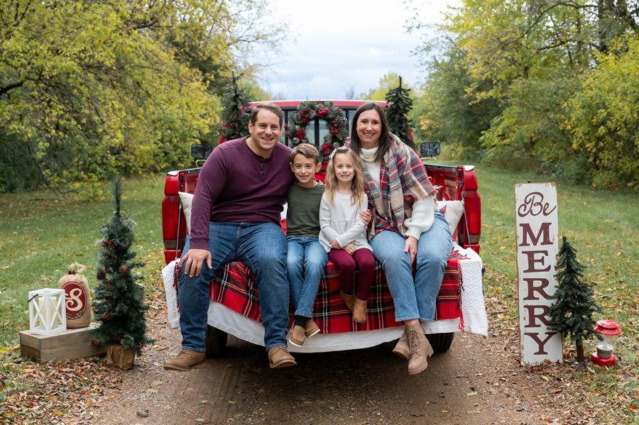 A family poses together in a red pickup truck with Christmas decorations and a plaid blanket.