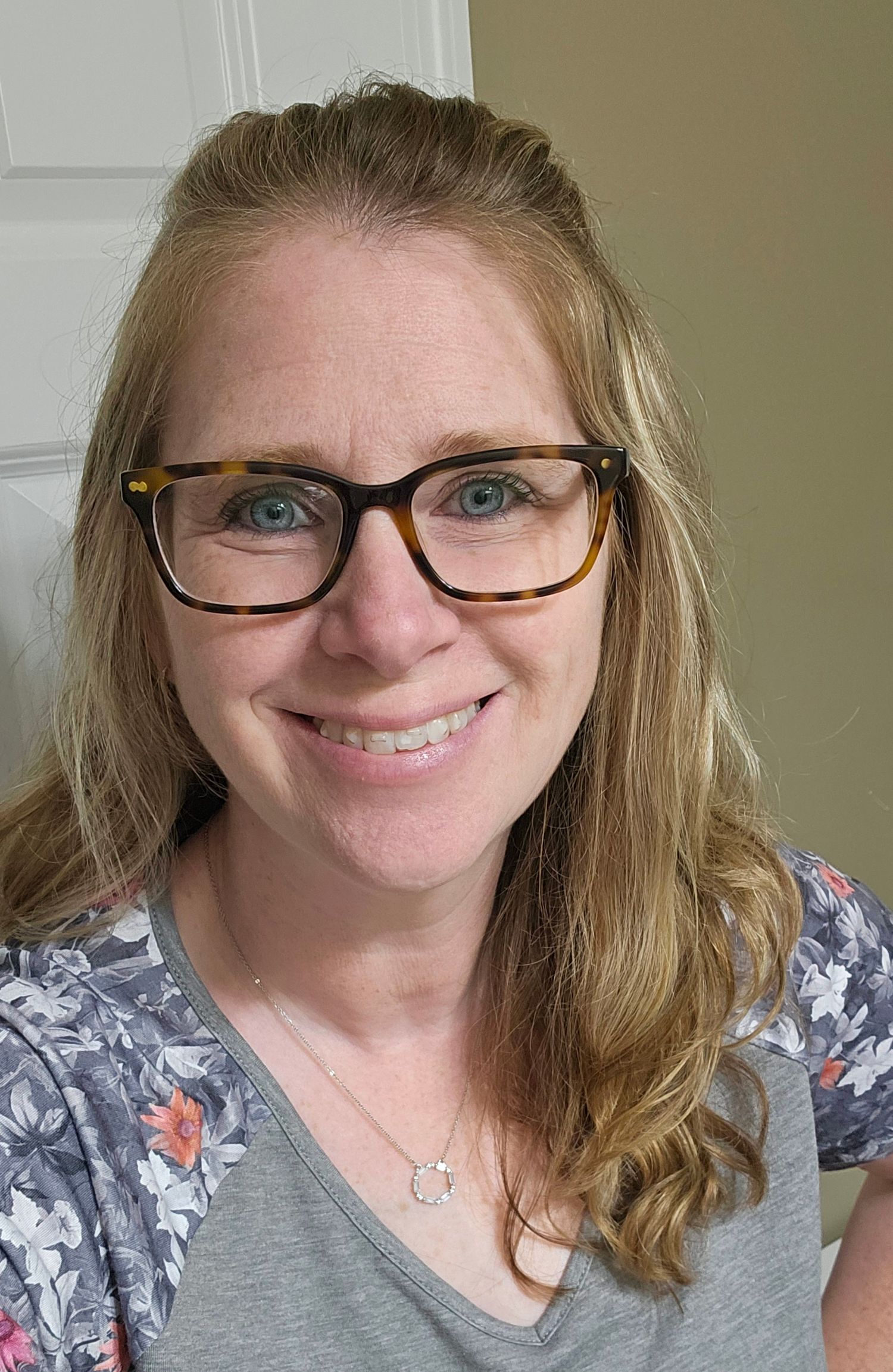 Smiling woman with blue eyes, tortoiseshell glasses, and blonde hair wearing a floral shirt and delicate necklace.