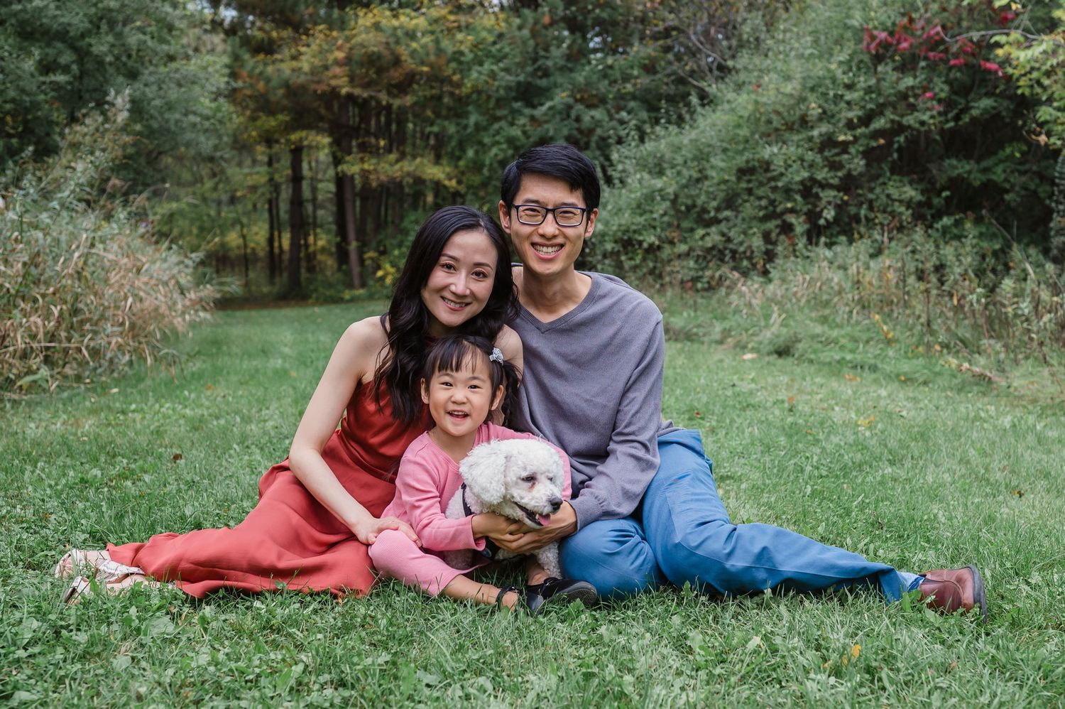 Family of three sitting on grass with small white dog during outdoor portrait session.