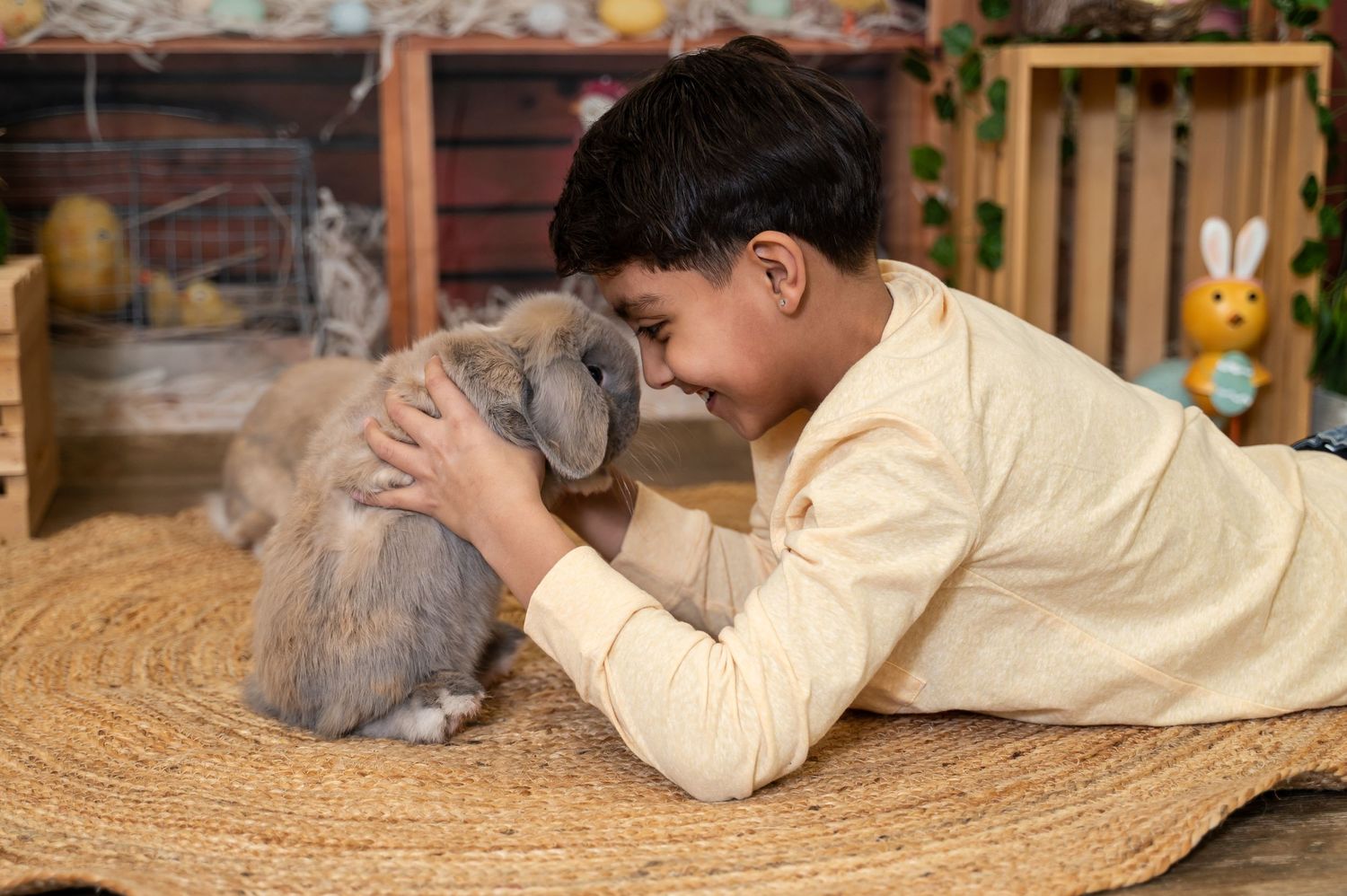A young person in a cream sweater lies on a round jute rug while playing with a fluffy gray bunny rabbit.