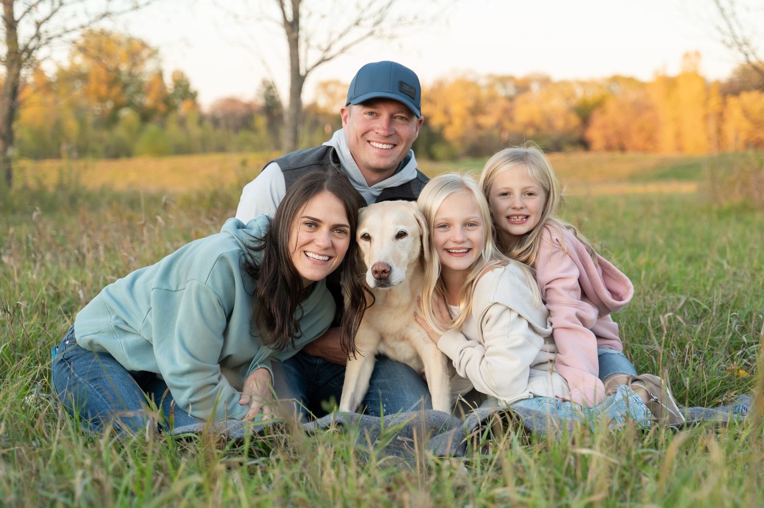 Family of four cuddling with their golden retriever dog in sunny autumn field.