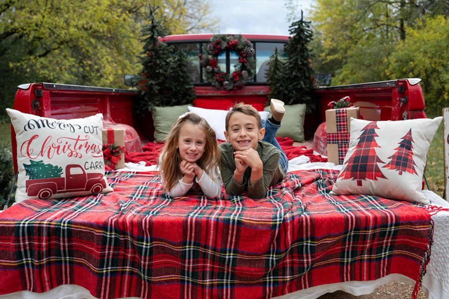 Two children lie on a plaid blanket in a truck bed decorated with Christmas pillows and trees.