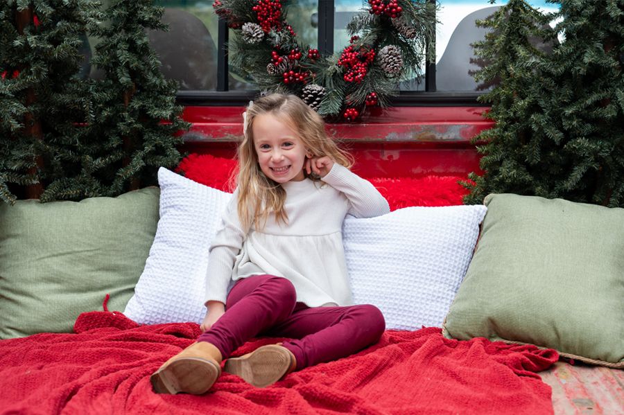 A child sits among white pillows on a red blanket in a Christmas-themed truck bed setting.