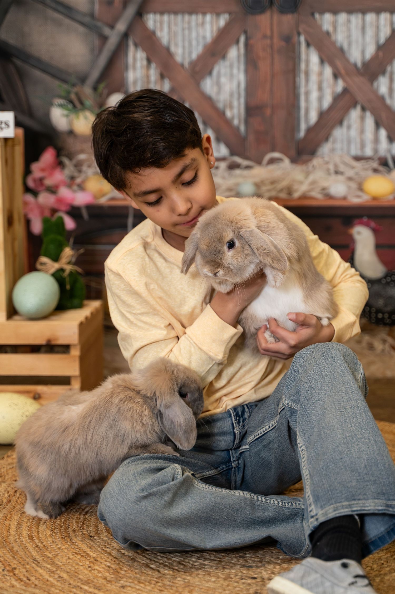 A young child in jeans and yellow shirt cuddles with a fluffy gray rabbit on a woven rug.