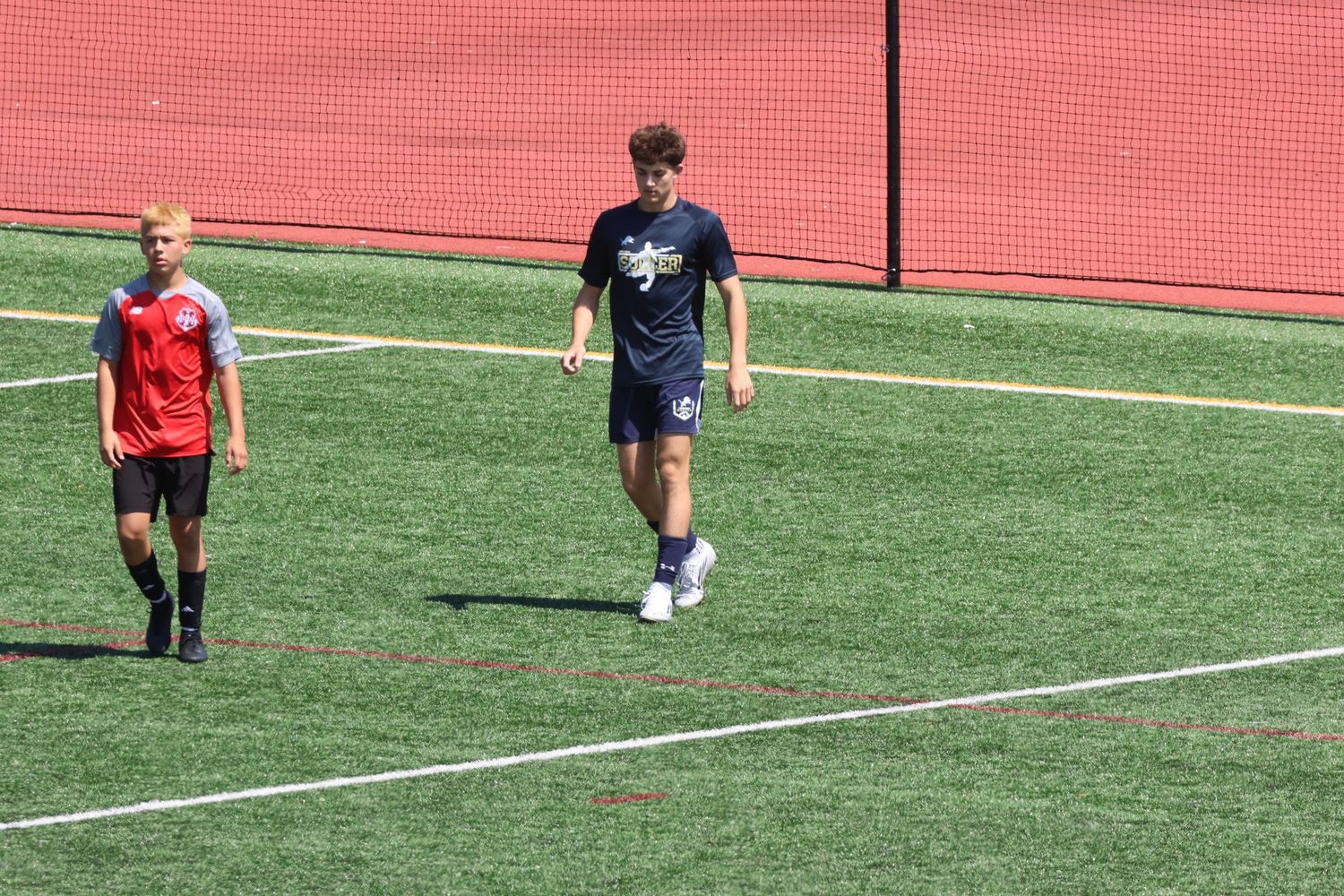 Two young soccer players walk on a bright green turf field during an outdoor summer soccer camp.