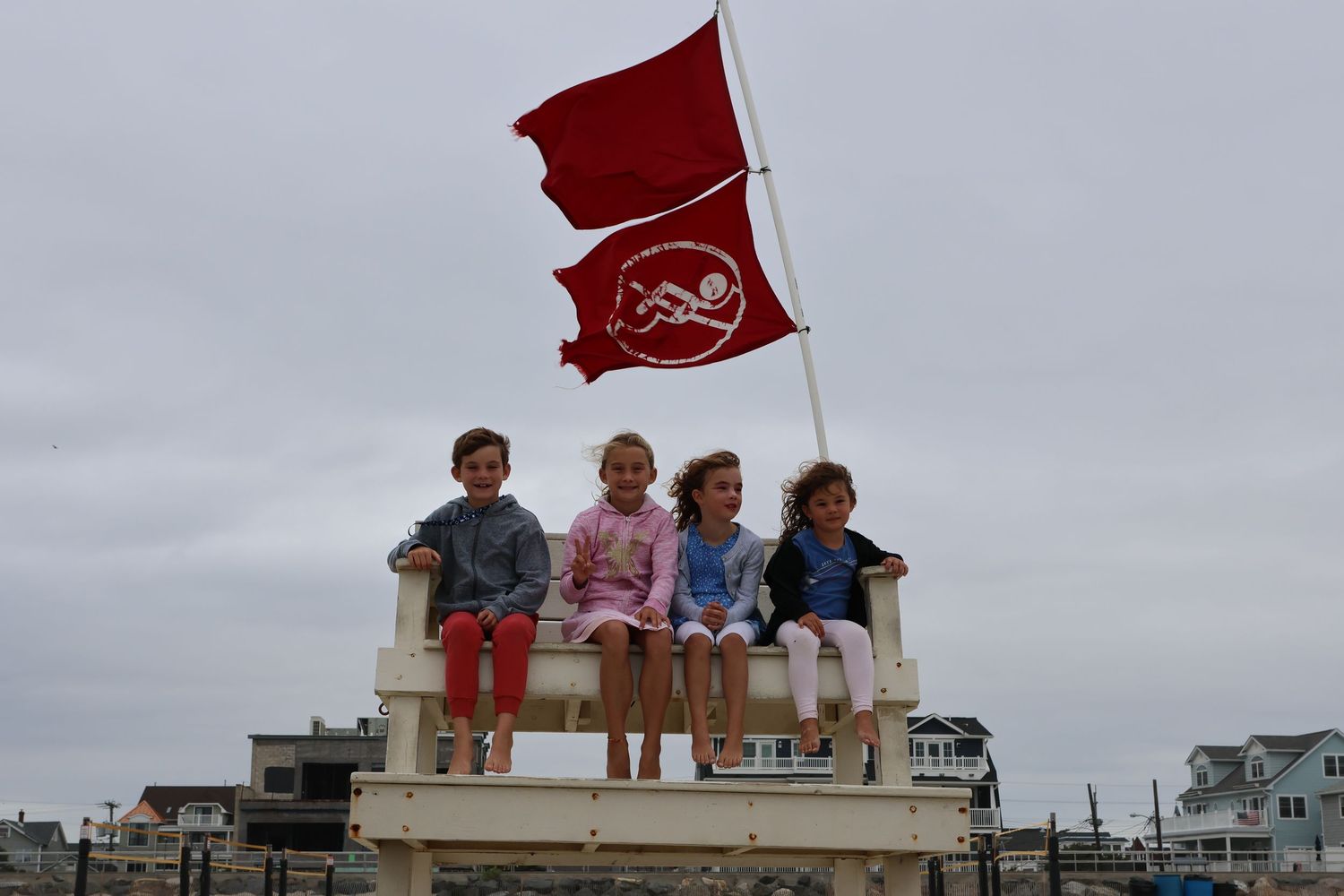 Four children sitting on a lifeguard stand with a red warning flag on a cloudy beach day.