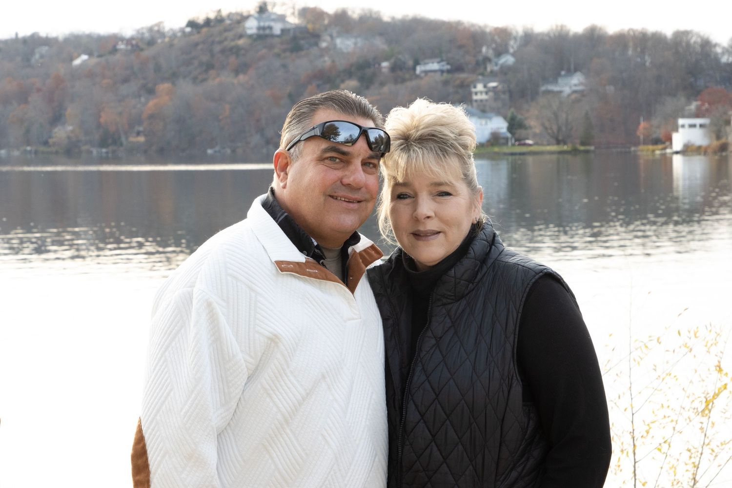 Smiling couple posing together near a lake with colorful autumn trees and hills in the background.