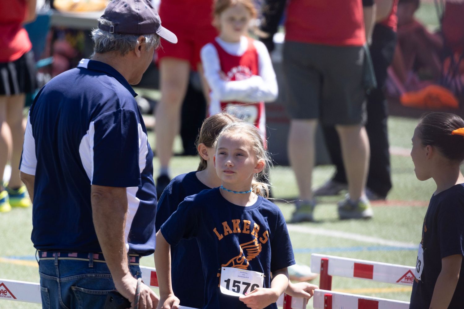 A young girl wearing a Larens jersey with bib number 1576 talks to a coach at a track meet.