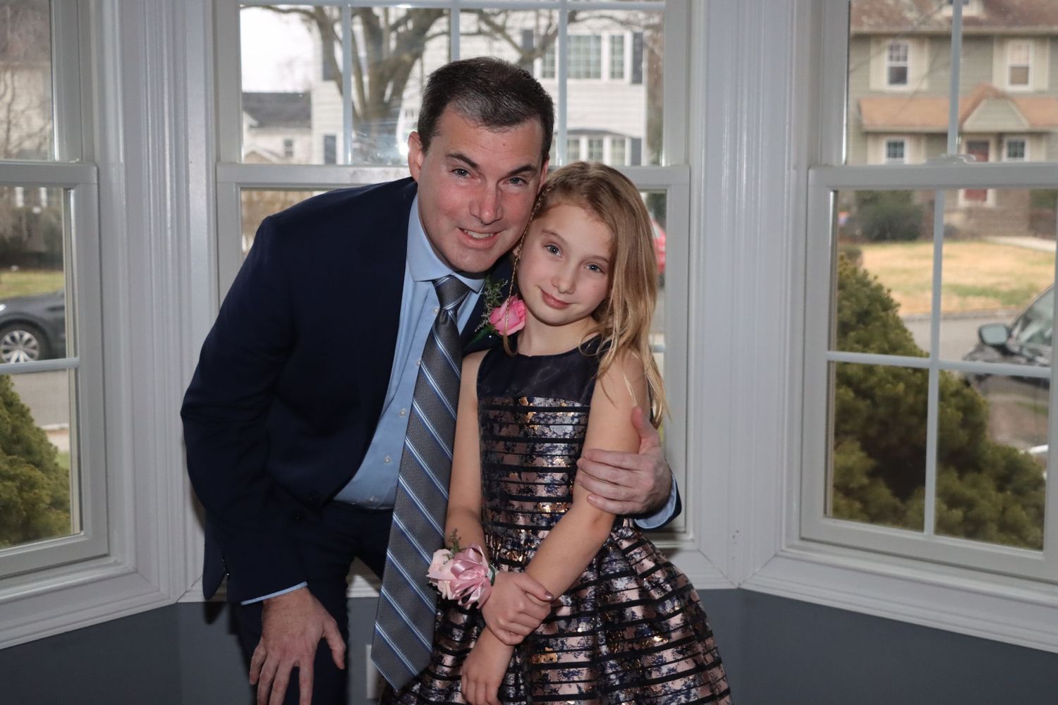 Man in a suit crouching beside a young girl in a sparkly dress posing near bright windows.