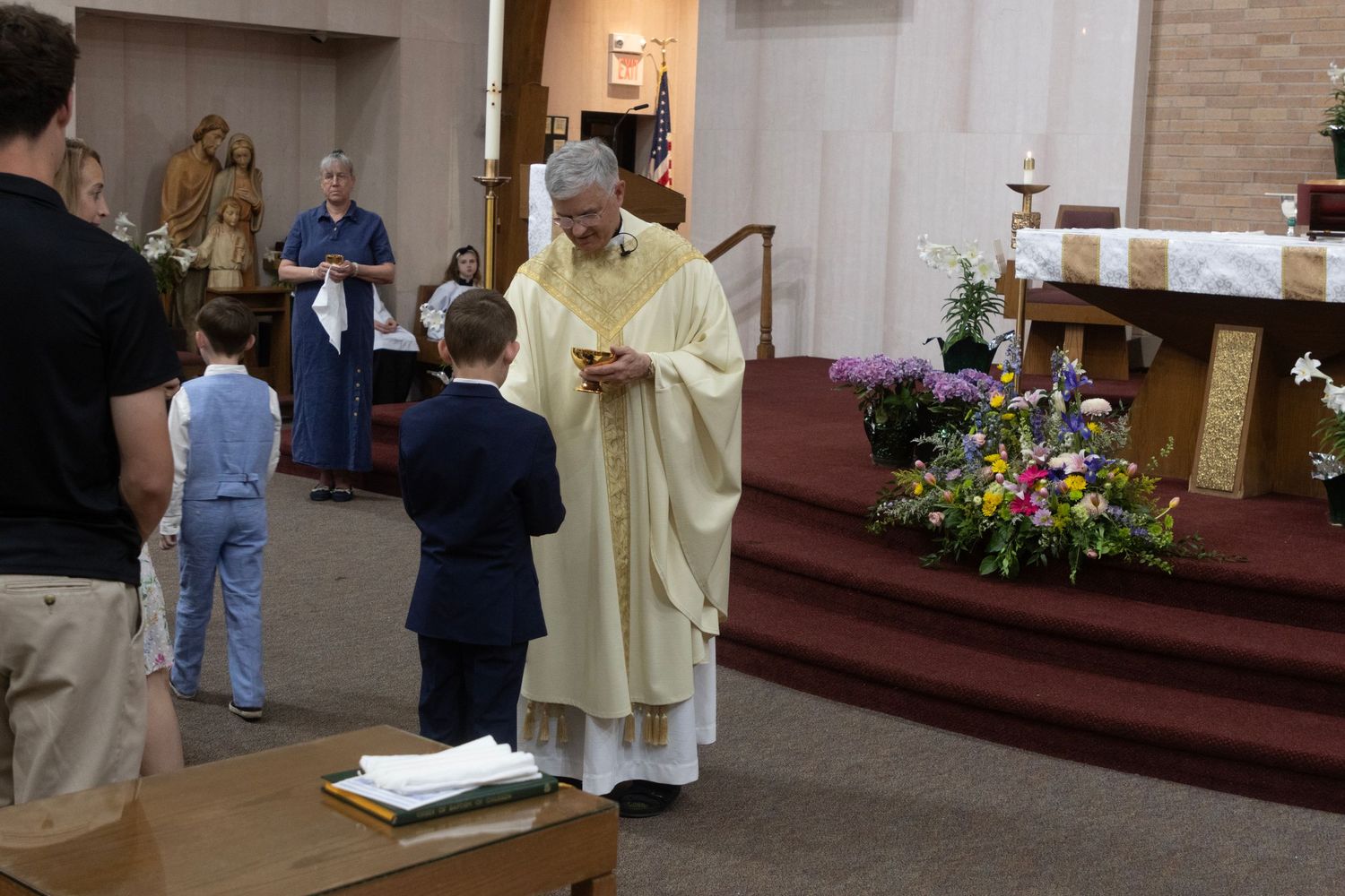 A priest in white vestments gives communion to a young boy in a suit during a Catholic church ceremony.