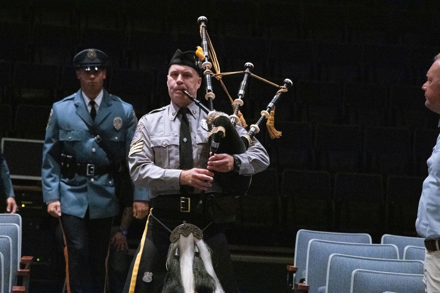 Uniformed bagpiper playing bagpipes while walking through auditorium rows with officers behind him.