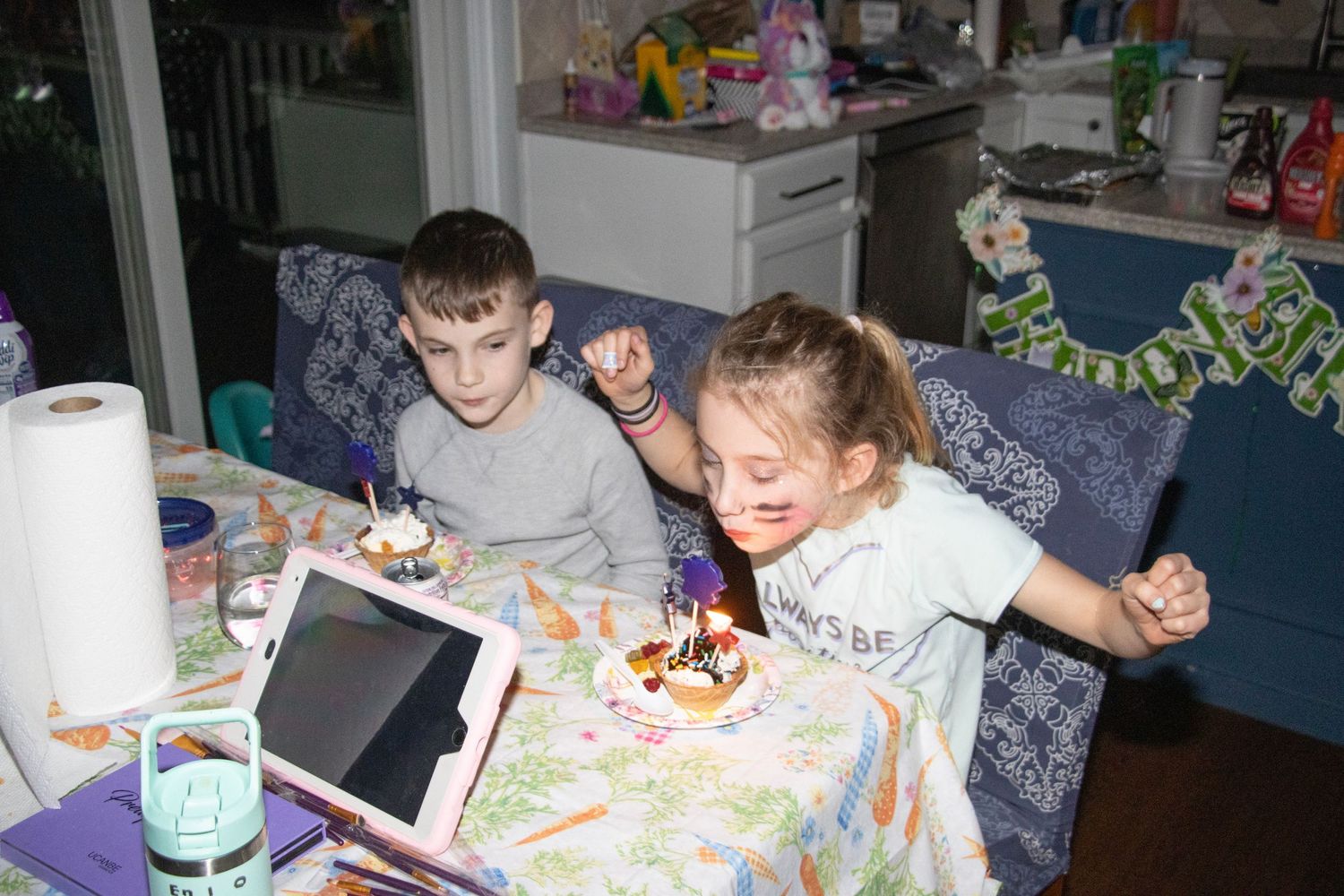Two children celebrating a birthday with a small cake and tablet on a colorful table.