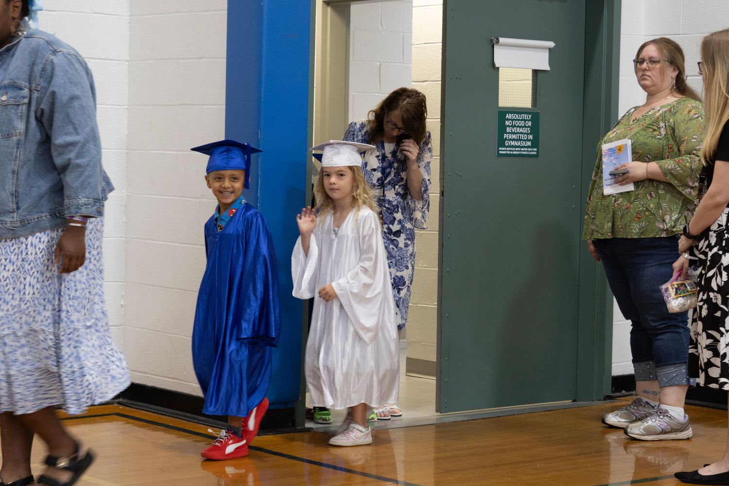 Young children in blue and white graduation caps and gowns walk into a gymnasium for a ceremony.