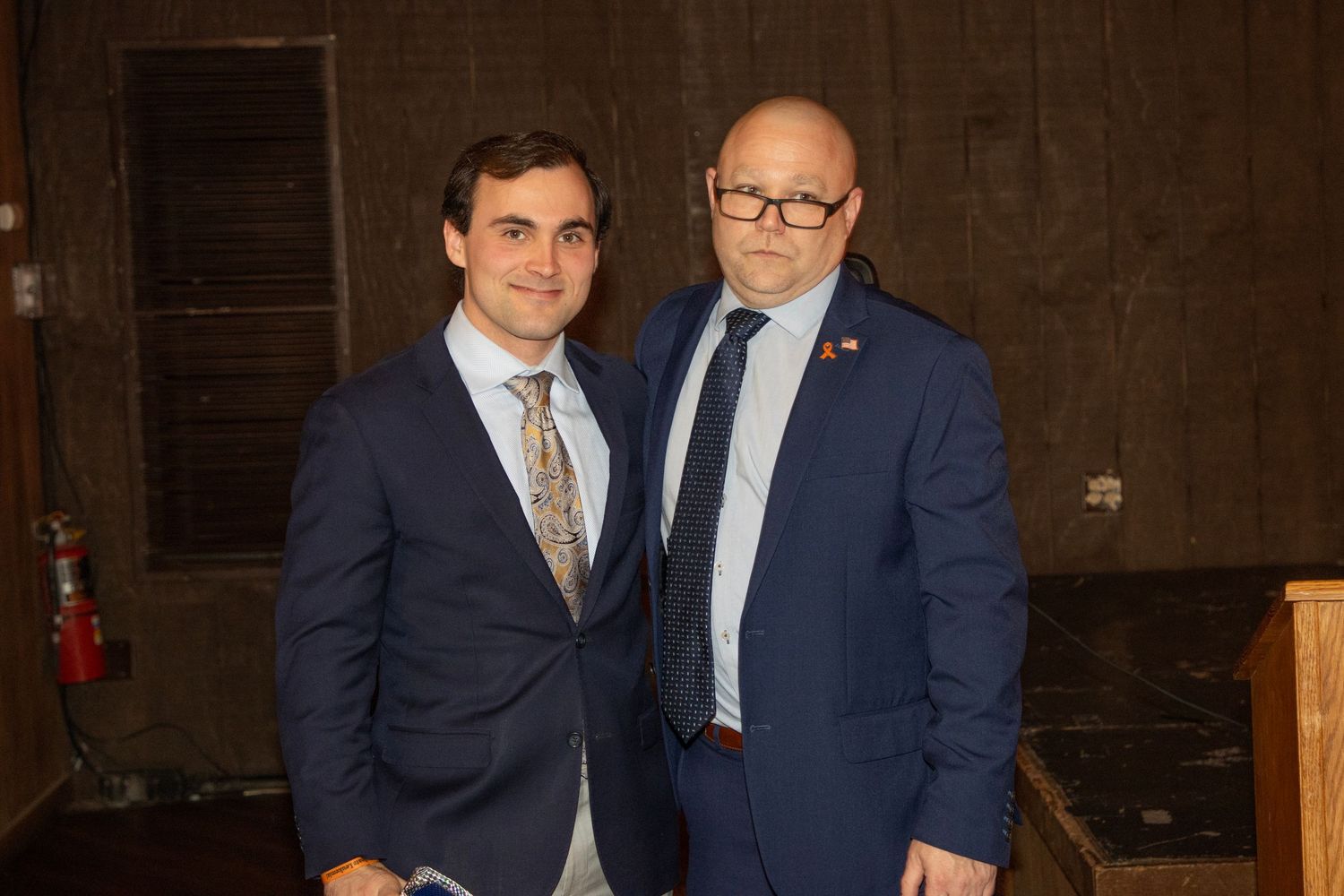 Two men in navy blue suits pose together smiling for a photo.