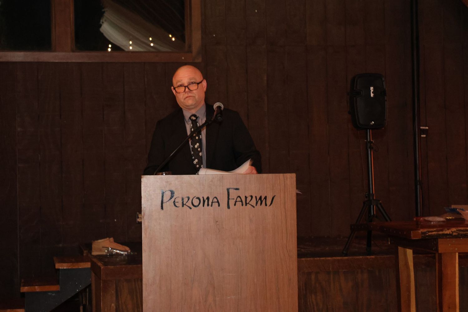 Person speaking at a Perona Farms wooden podium in a dimly lit venue.