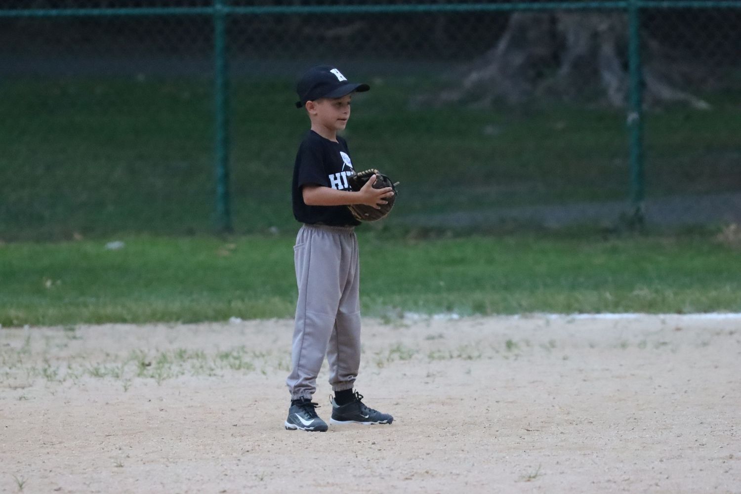 Young boy in baseball uniform standing on a dirt field holding a glove during a game.