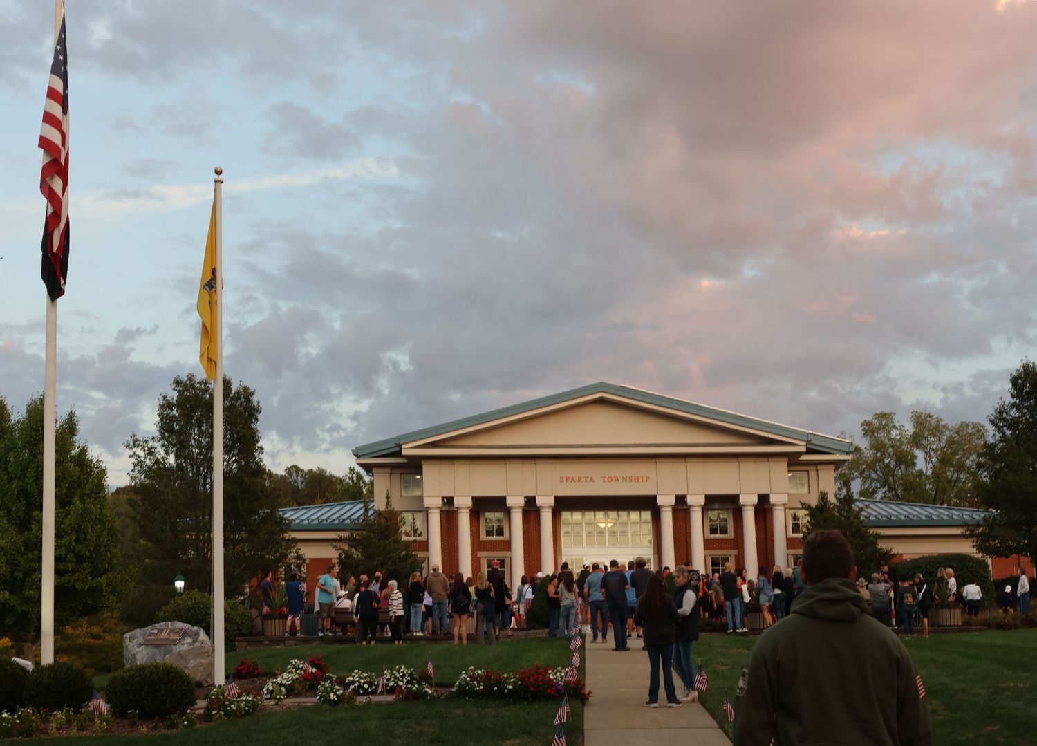 Crowd gathers outside a large building at dusk with American and state flags flying.