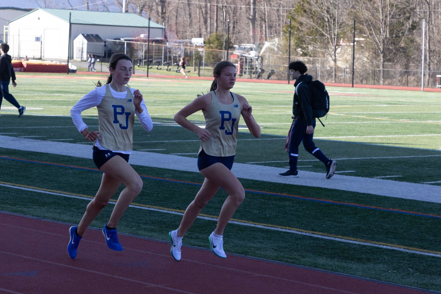 Two female athletes in matching uniforms run together on an outdoor track during a meet.