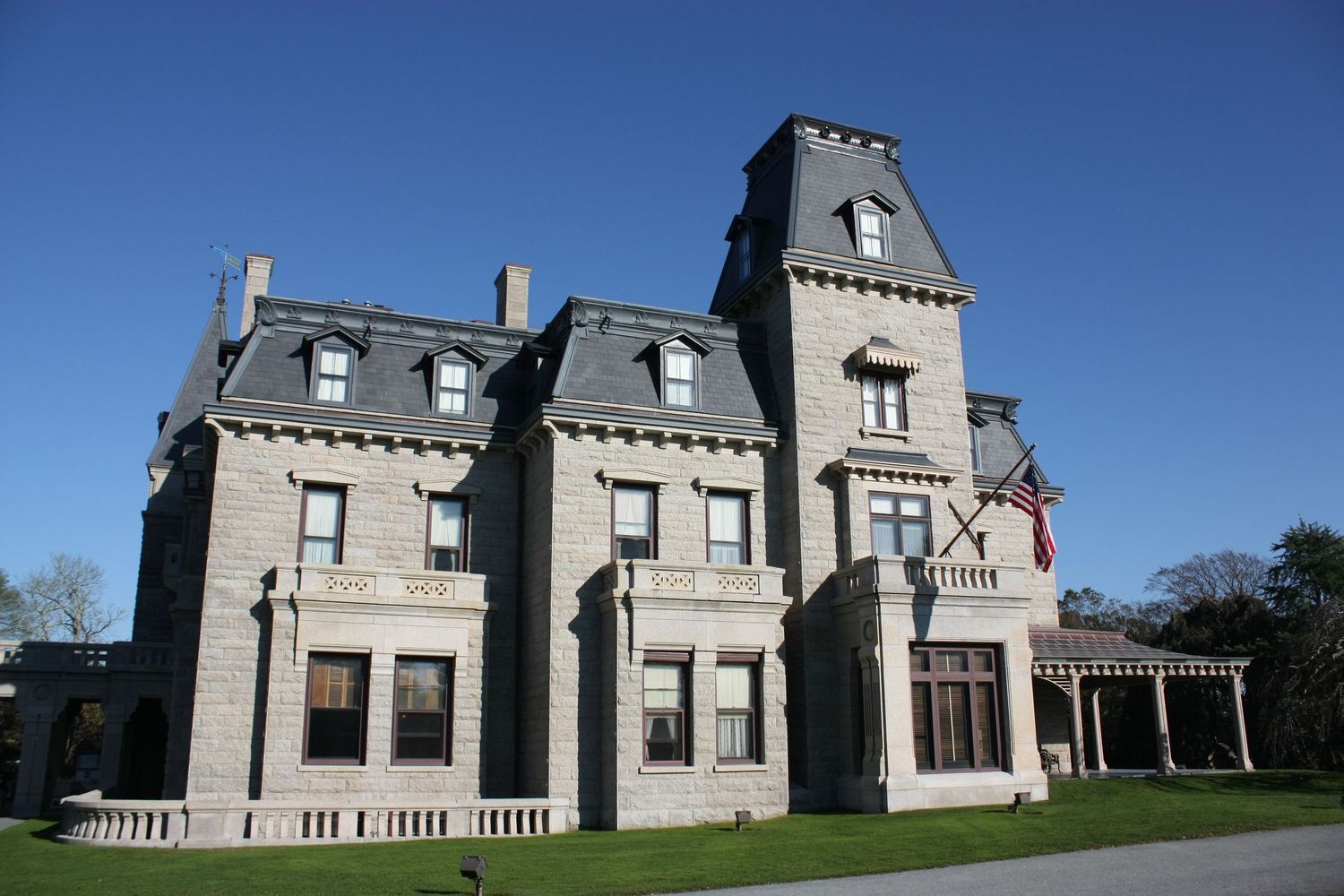 Grand stone mansion with French chateau-style architecture featuring dormer windows and slate roof under clear blue sky.
