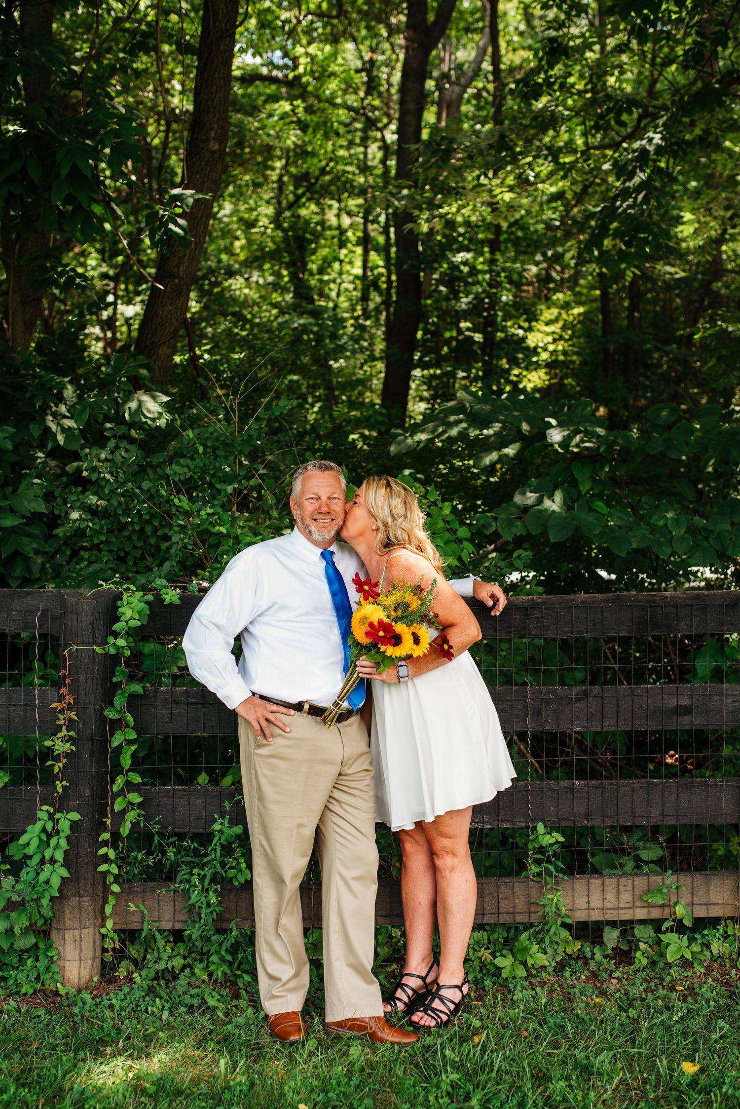 A couple poses together against a rustic wooden fence surrounded by lush green forest while holding a vibrant sunflower bouquet.
