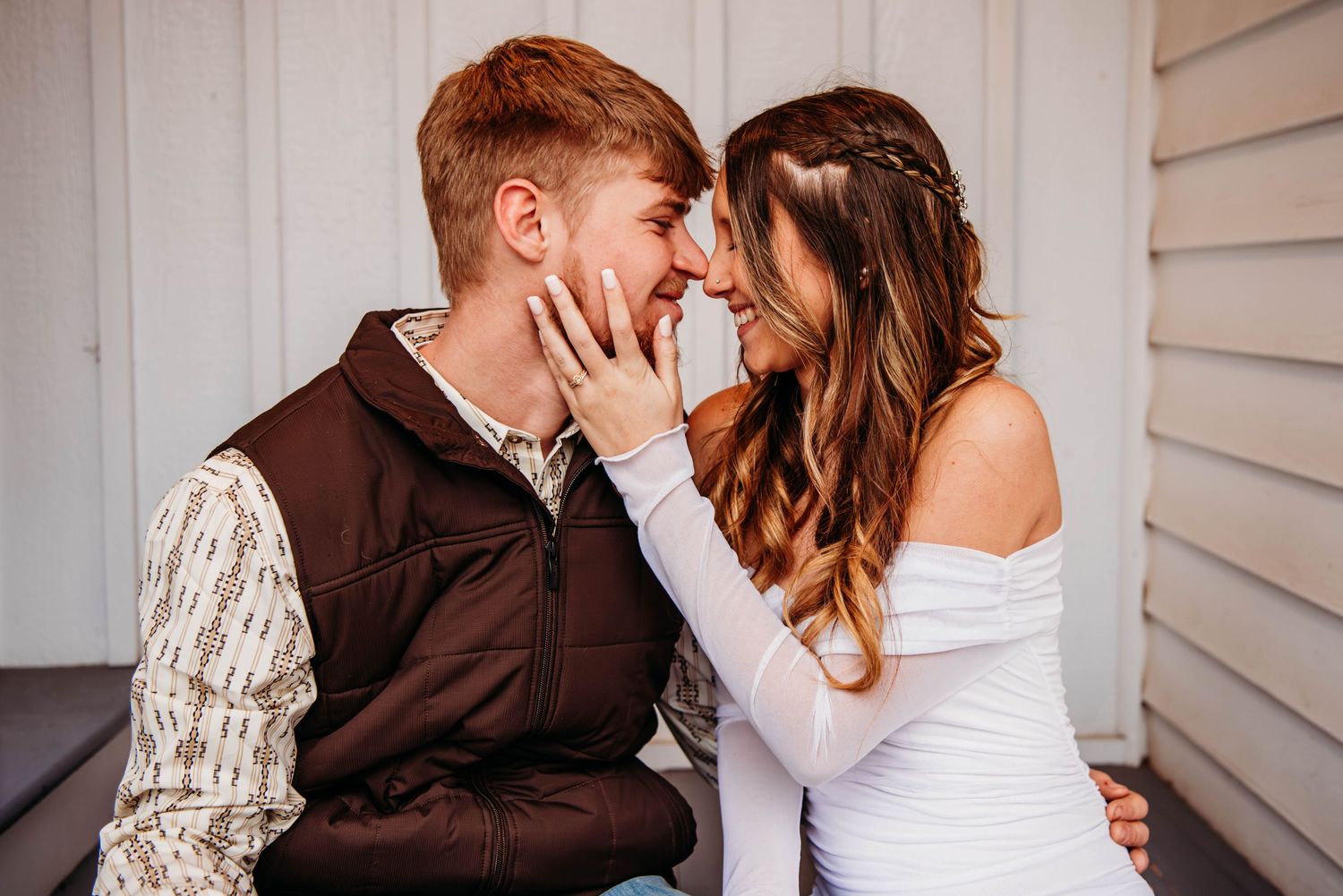 A young couple shares a romantic moment, noses touching, as the woman gently holds the man's face outdoors.