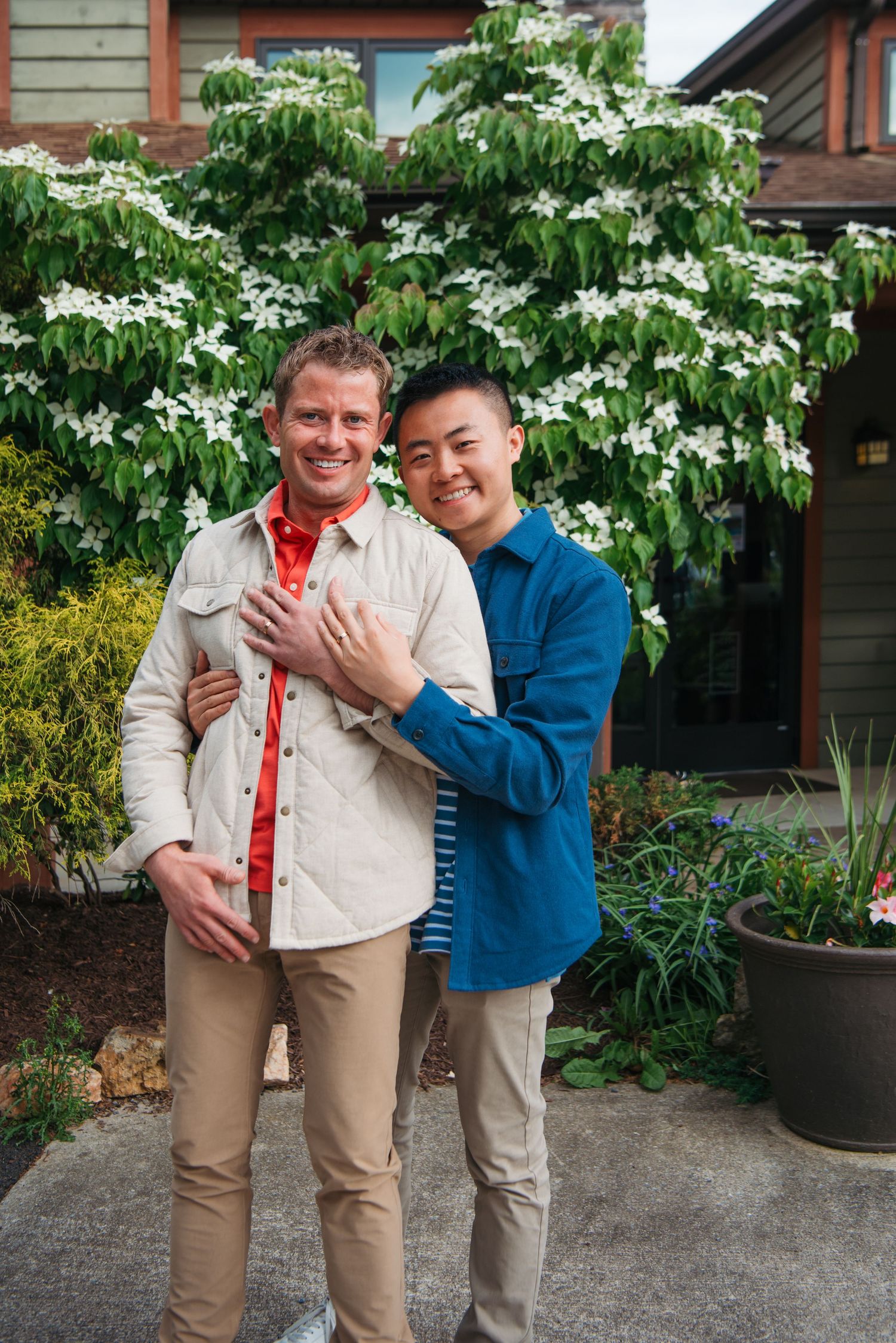 Two smiling men pose together outdoors in front of a flowering dogwood tree near a house.
