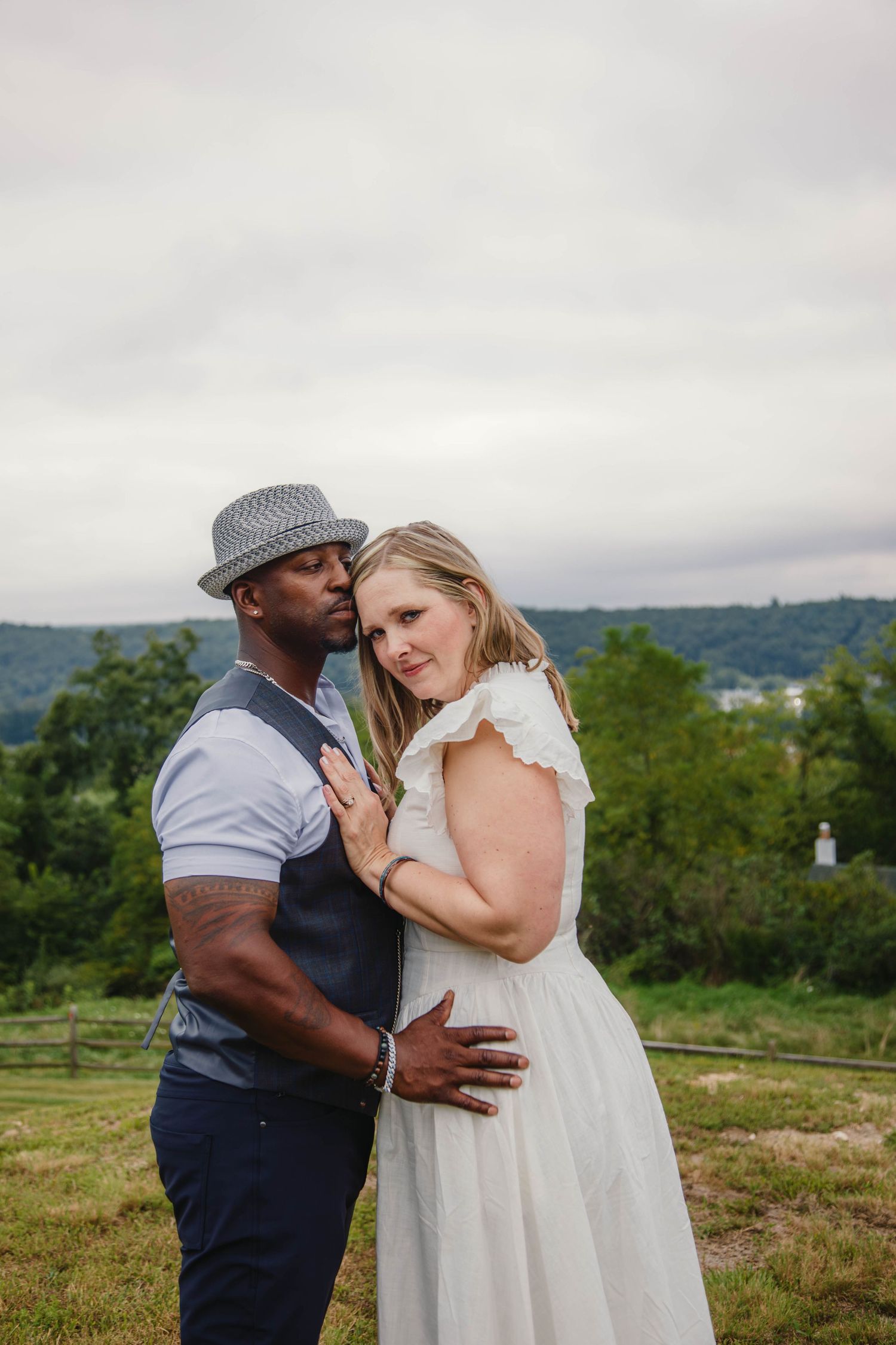 Interracial couple embracing outdoors in wedding attire, man in gray hat and vest, woman in white dress, green hills behind.