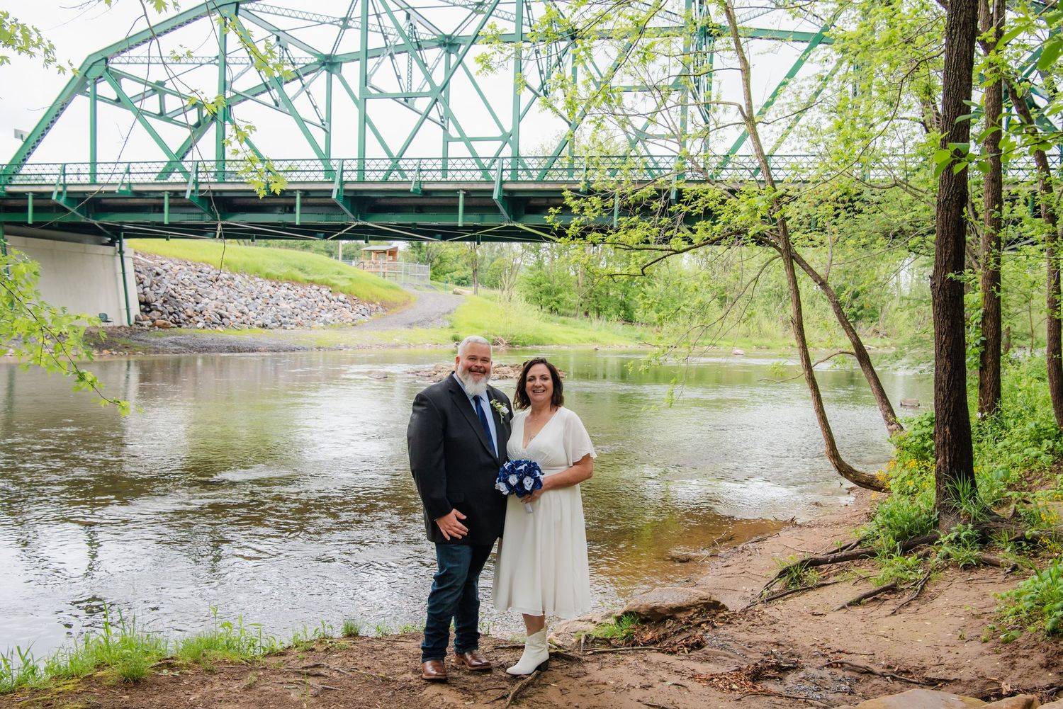 Couple poses by a river with a green steel bridge backdrop, bride in white dress holding a blue bouquet.