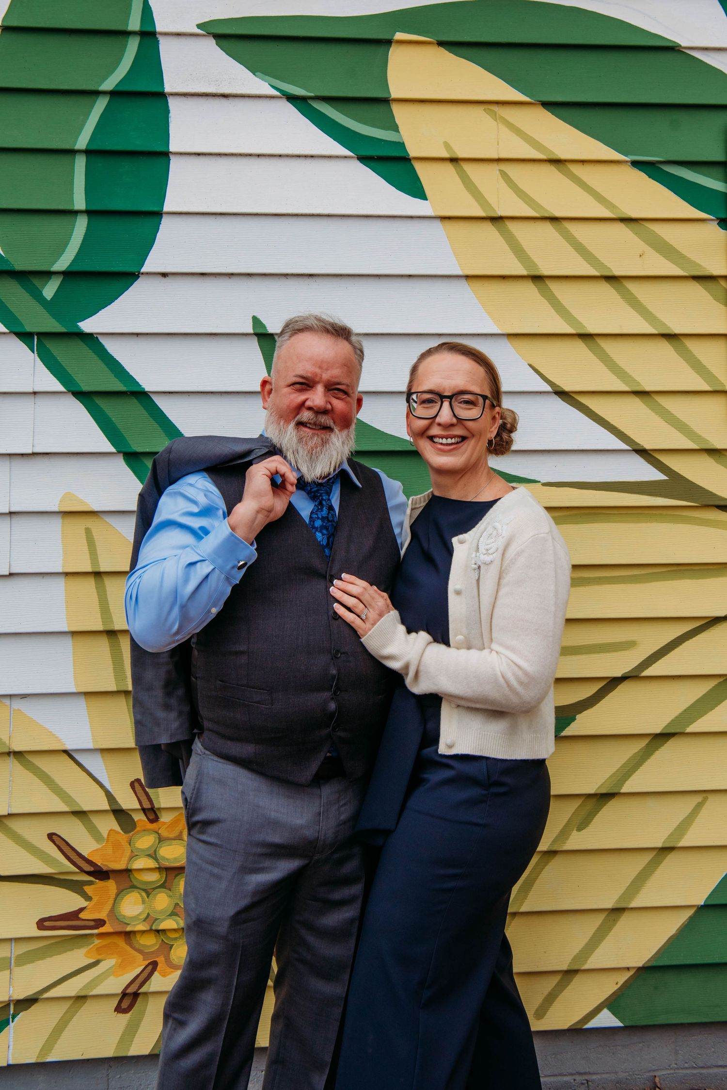 Smiling couple posing in front of colorful floral mural, man in gray vest and woman in navy dress with white cardigan.