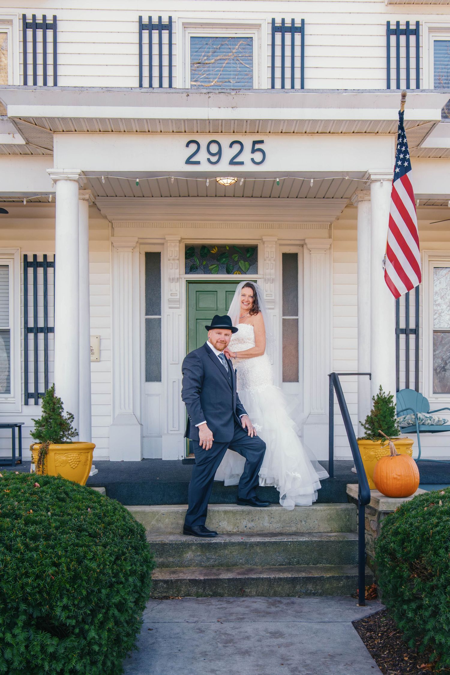 Bride and groom pose on steps of white house numbered 2925 with American flag, pumpkin decor, in wedding attire.