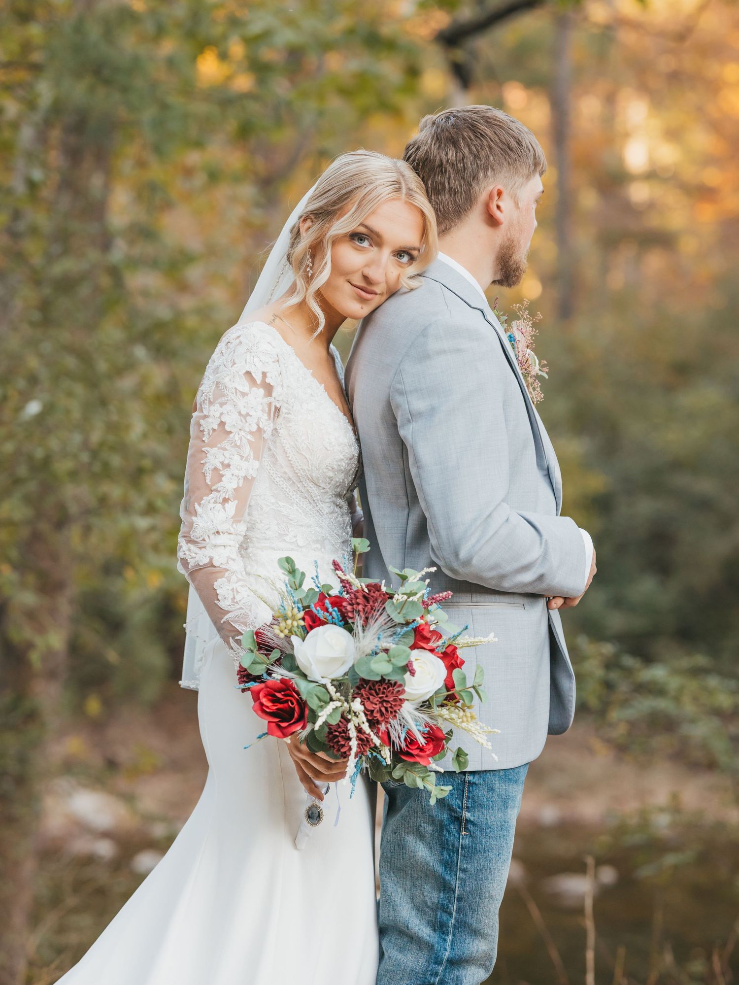 Bride in lace gown holds red and white bouquet, leaning on groom in gray blazer and jeans outdoors.