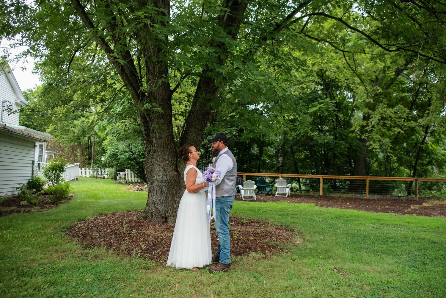 A bride and groom share a romantic moment under a large tree in a lush green backyard on their wedding day.