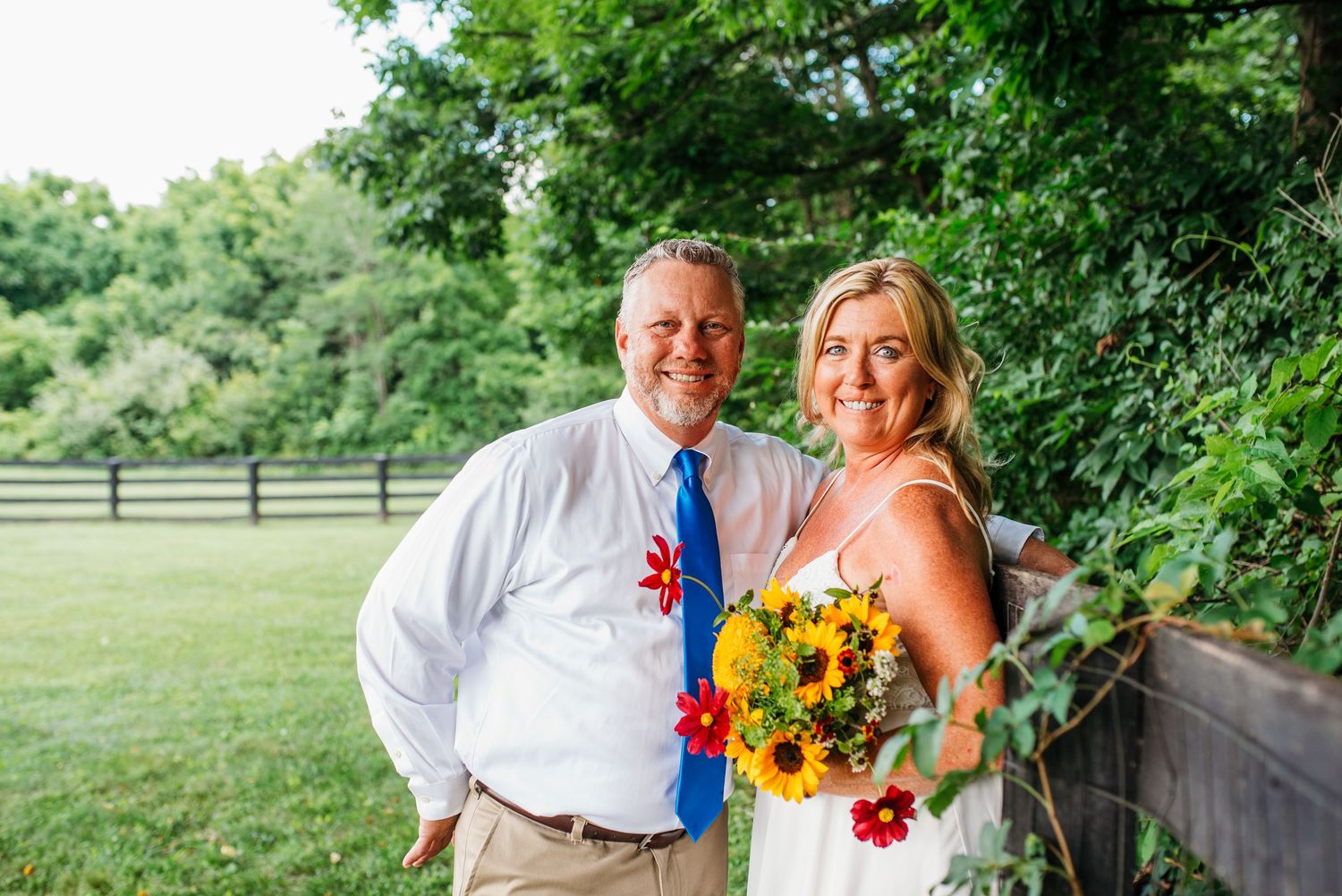 Smiling couple posing outdoors at a wedding, woman holding colorful sunflower bouquet, lush green trees in background.