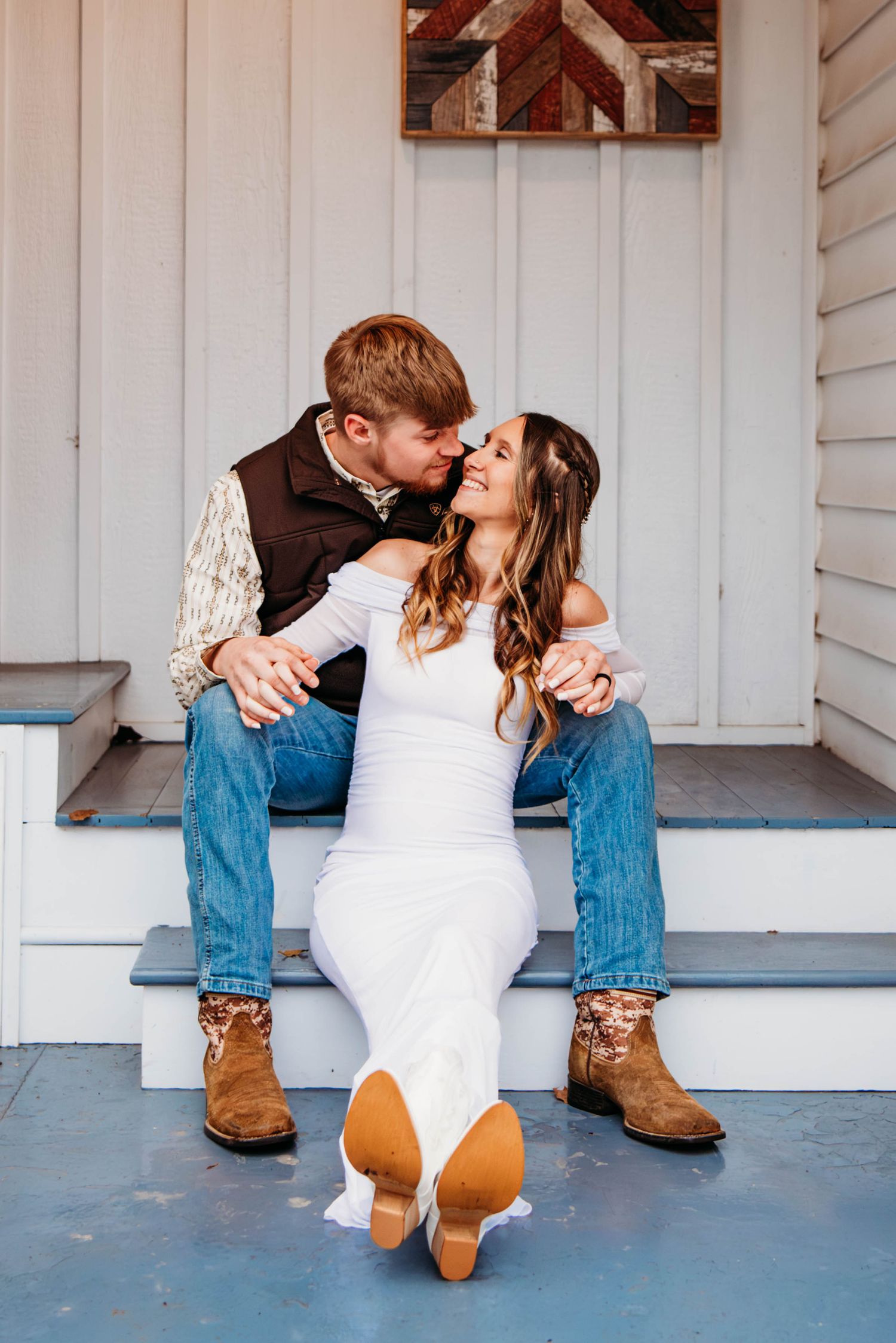 Couple in jeans and cowboy boots sitting on porch steps, sharing a romantic kiss in rustic wedding attire.