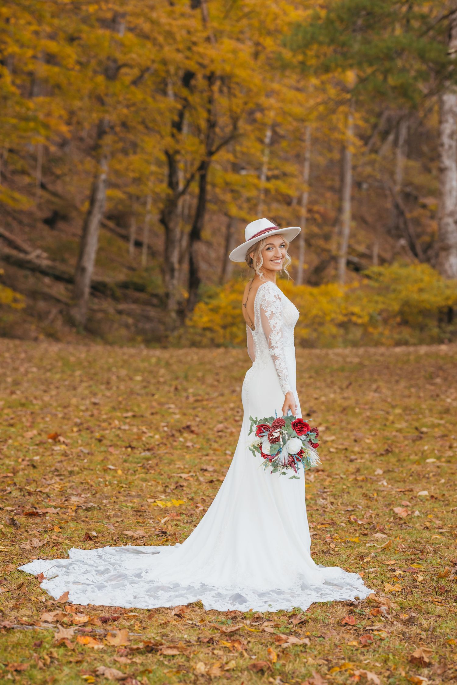 Bride in white lace gown with hat holds red bouquet among autumn foliage in a serene outdoor setting.
