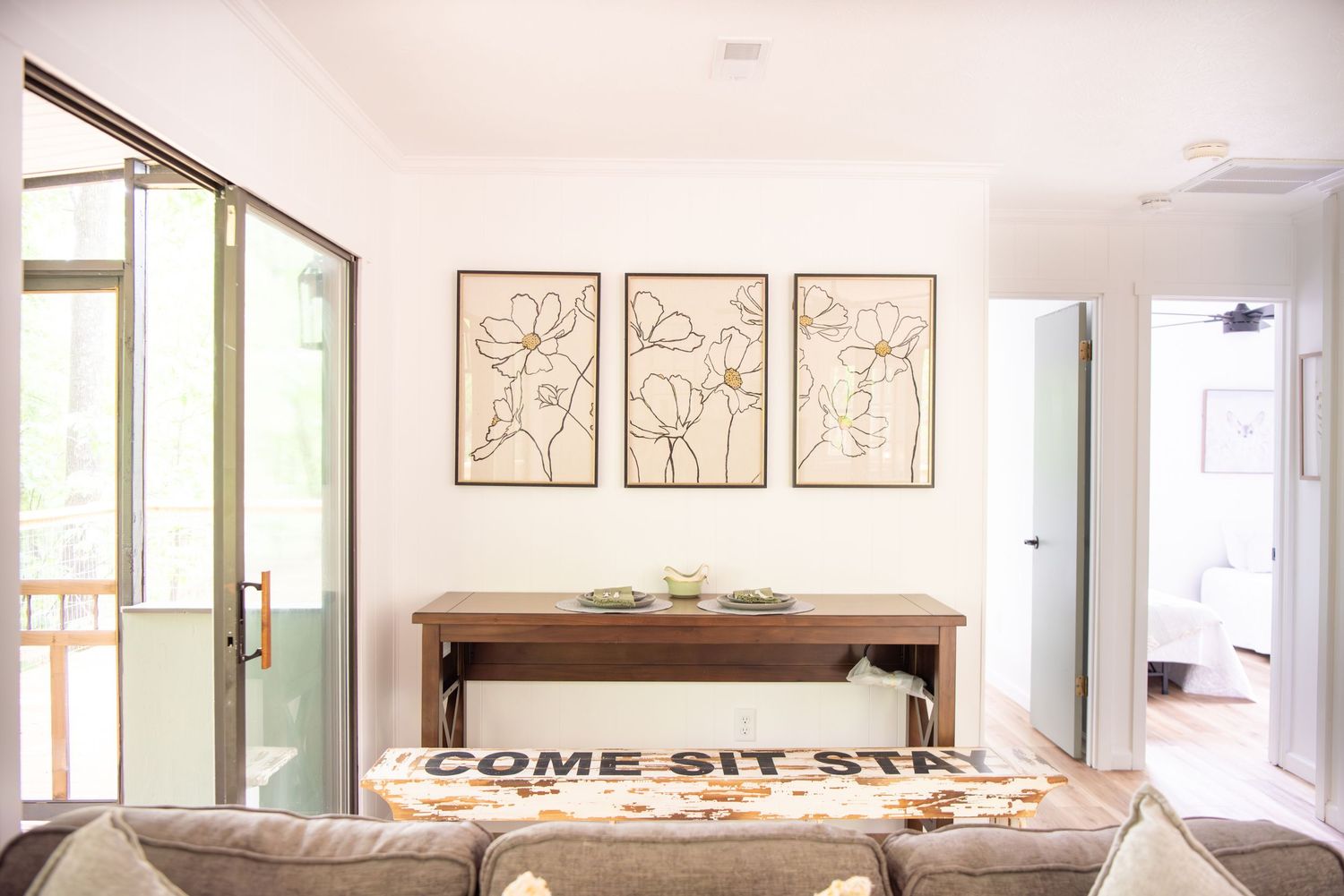 Modern minimalist dining room with three framed botanical prints and 'Come Sit' text on table.