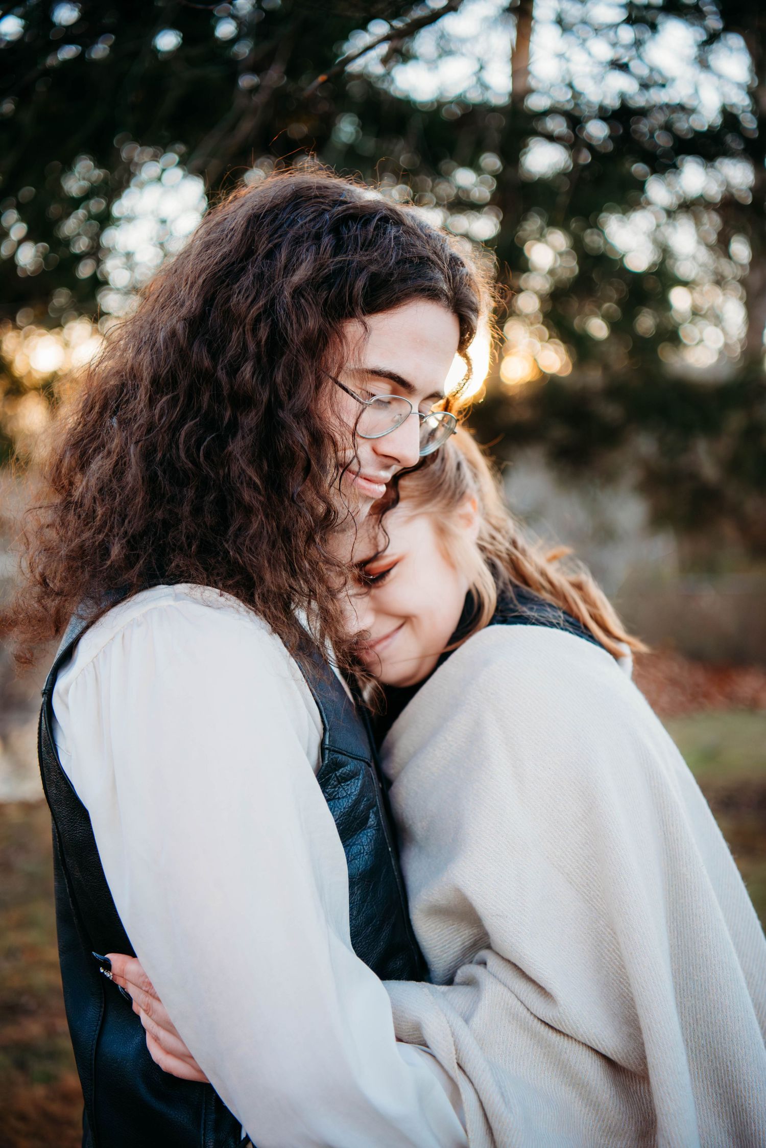 Two people share a warm embrace outdoors at golden hour, one with curly hair and glasses smiling tenderly.