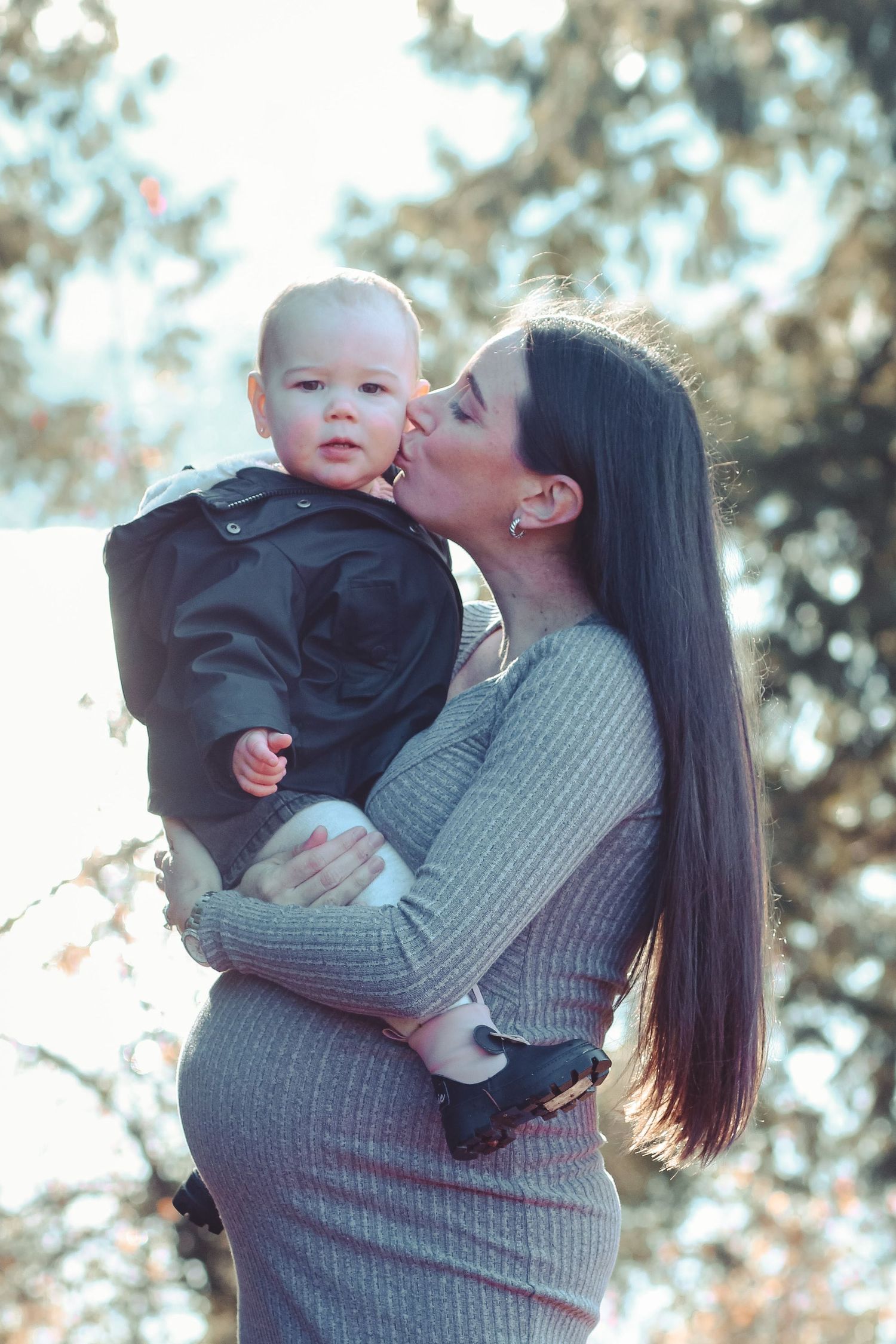 Outdoors maternity photo session winter time with toddler. Mom is holding toddler above her baby bump and giving her a gentle kiss on cheek.
