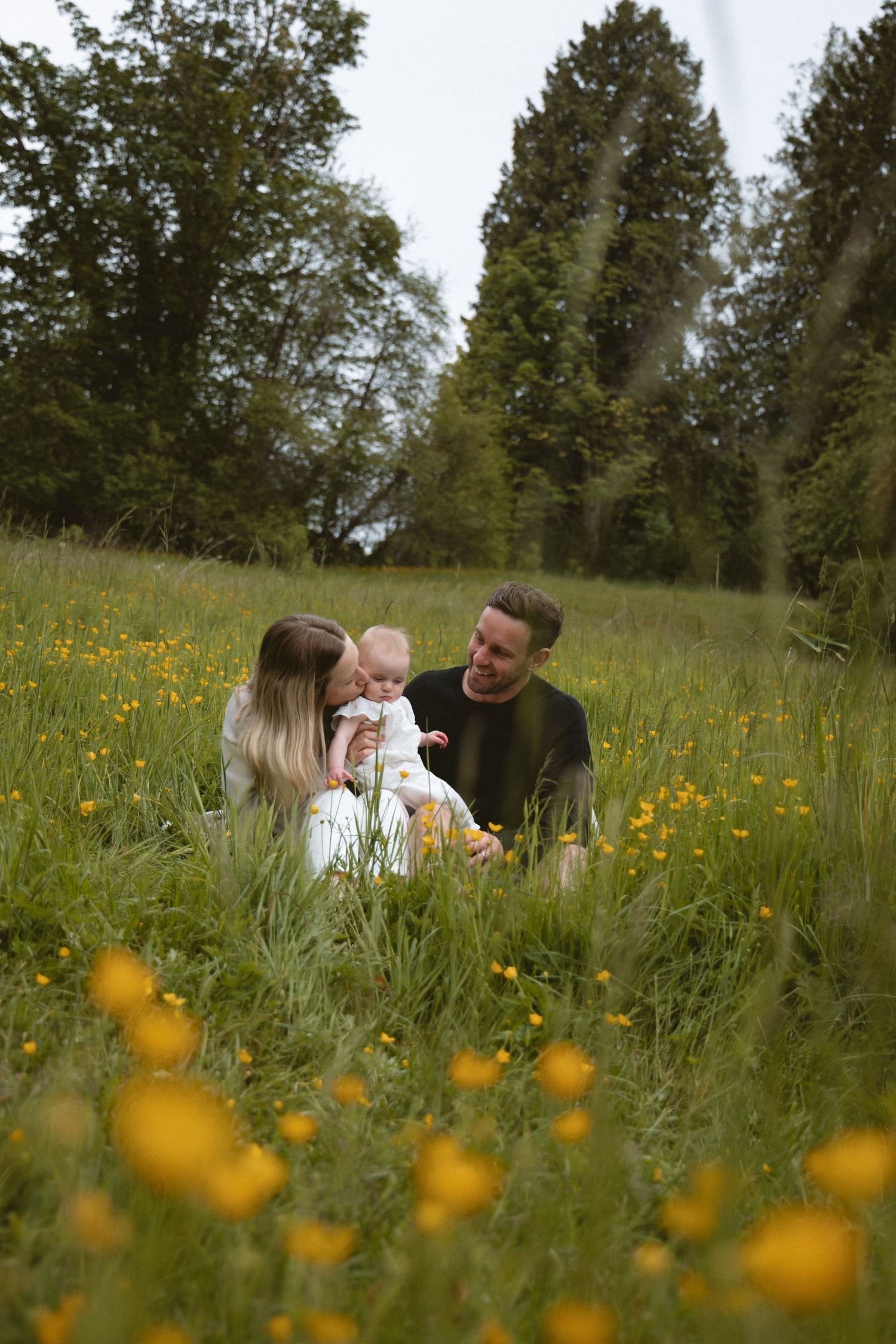 Candid family photoshoot with a couple and their baby in a field of flowers hugging the baby between them.