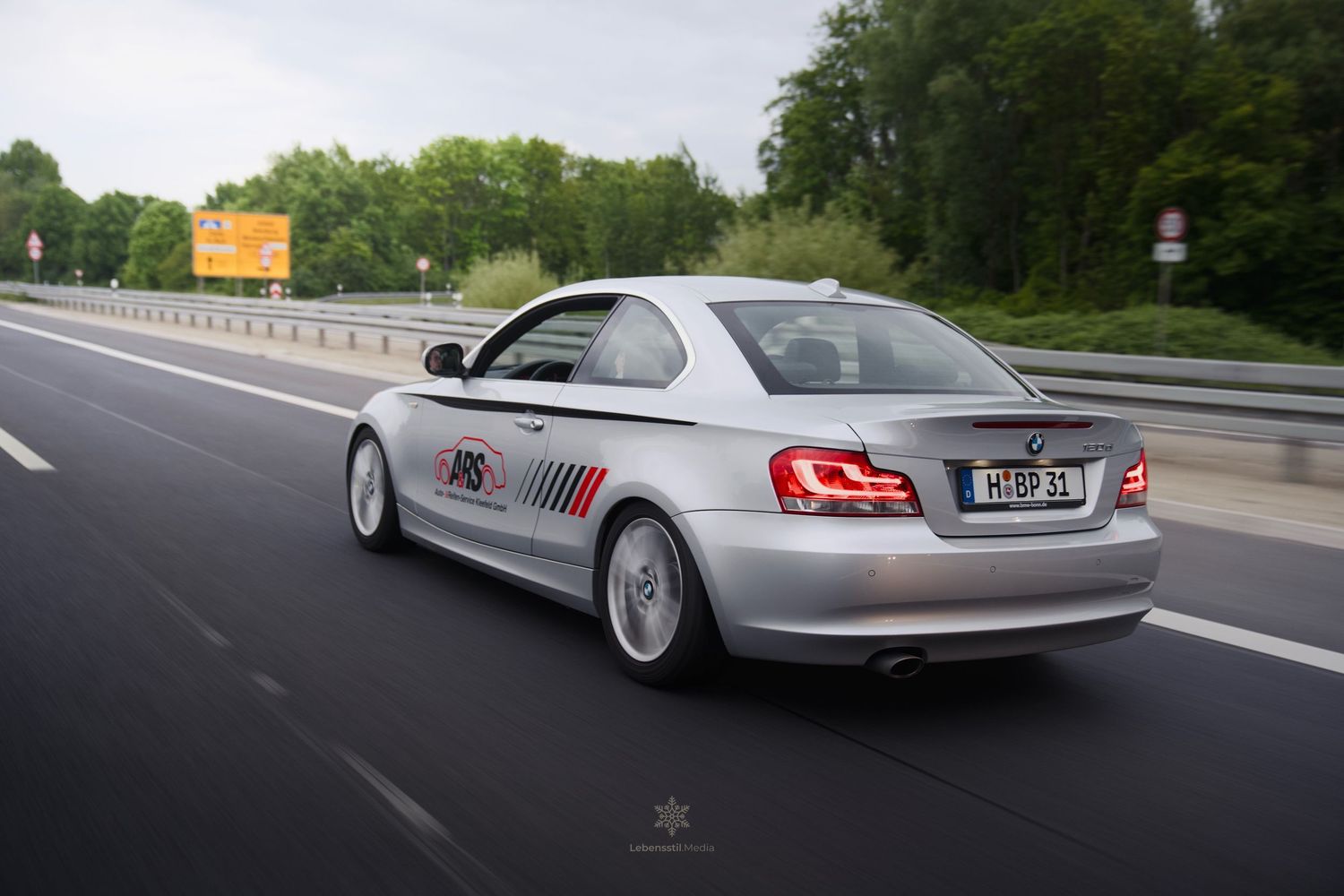 A silver luxury car with custom wheels driving on a highway during daytime.