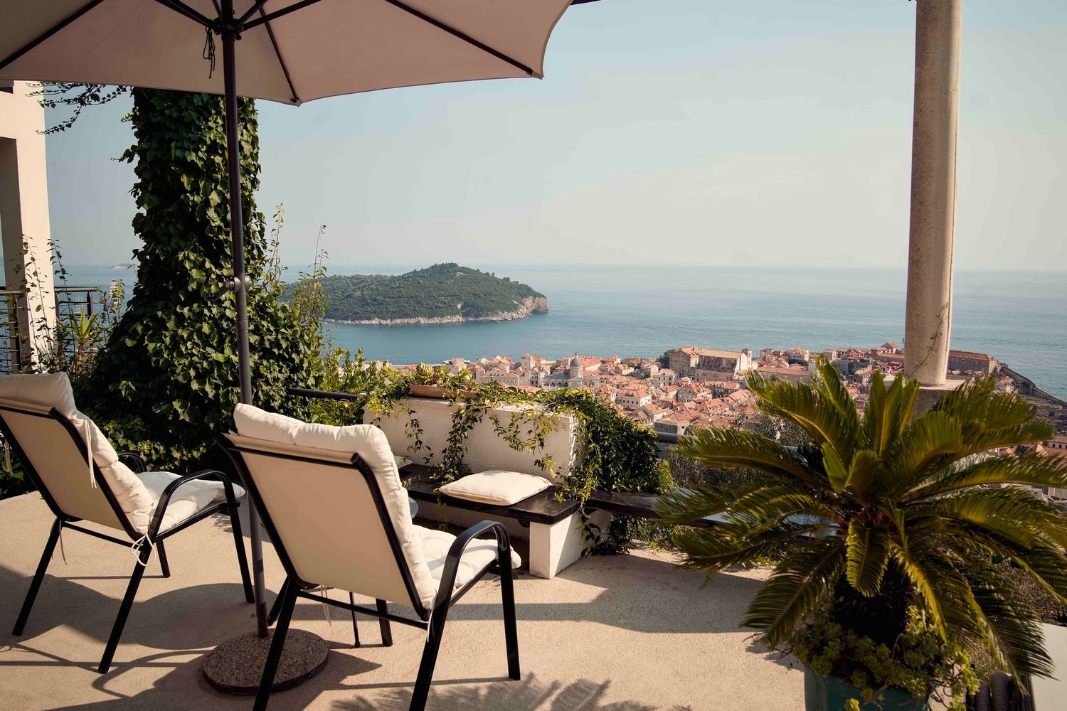 Outdoor terrace with patio furniture overlooking coastal Mediterranean town and sea with palm tree in foreground.