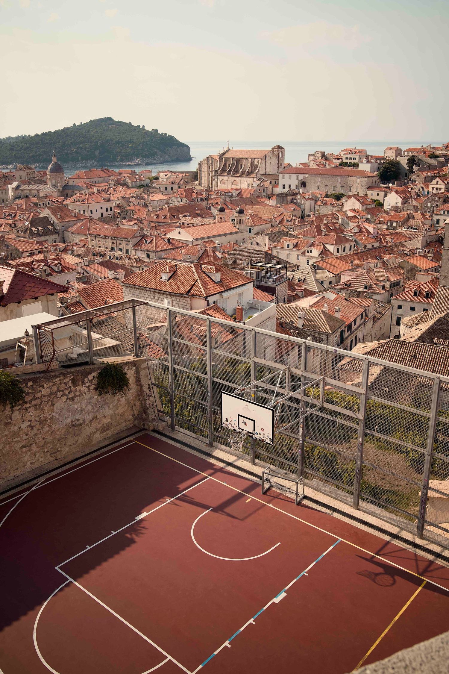 Aerial view of a rooftop basketball court overlooking historic Dubrovnik's terracotta rooftops and Adriatic coastline.